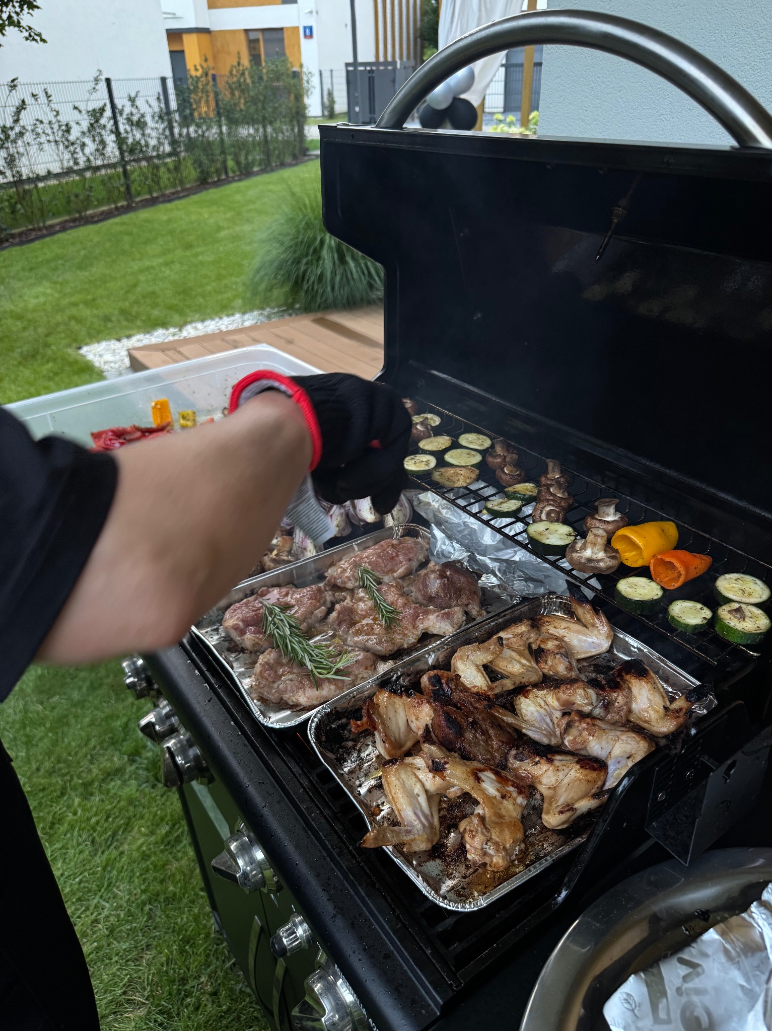 Przygotowywanie dań z grilla: mięso z rozmarynem, skrzydełka i warzywa (cukinia, papryka, pieczarki) na ruszcie. Osoba w rękawicy kulinarna doprawia potrawy.