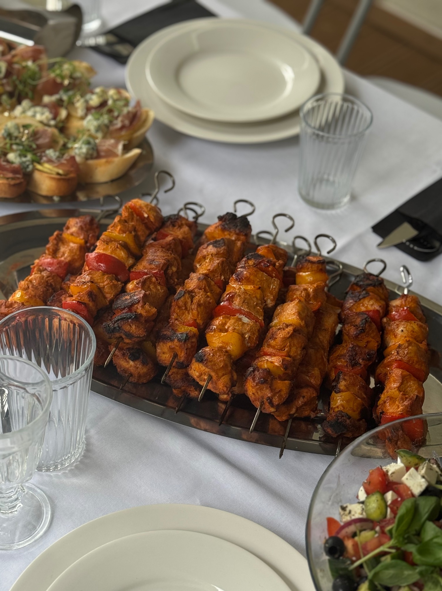 Szklane naczynia i srebrna taca z szaszłykami z kurczaka i warzyw, obok sałatka grecka i kanapki na białym obrusie. Widok z góry na stół cateringowy.