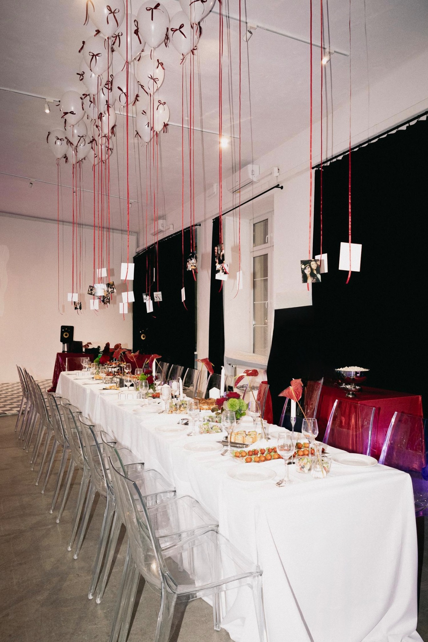 Uroczysty stół z białym obrusem, przezroczystymi krzesłami i dekoracjami: balony z czerwonymi wstążkami i zdjęciami. Przygotowany na imprezę.