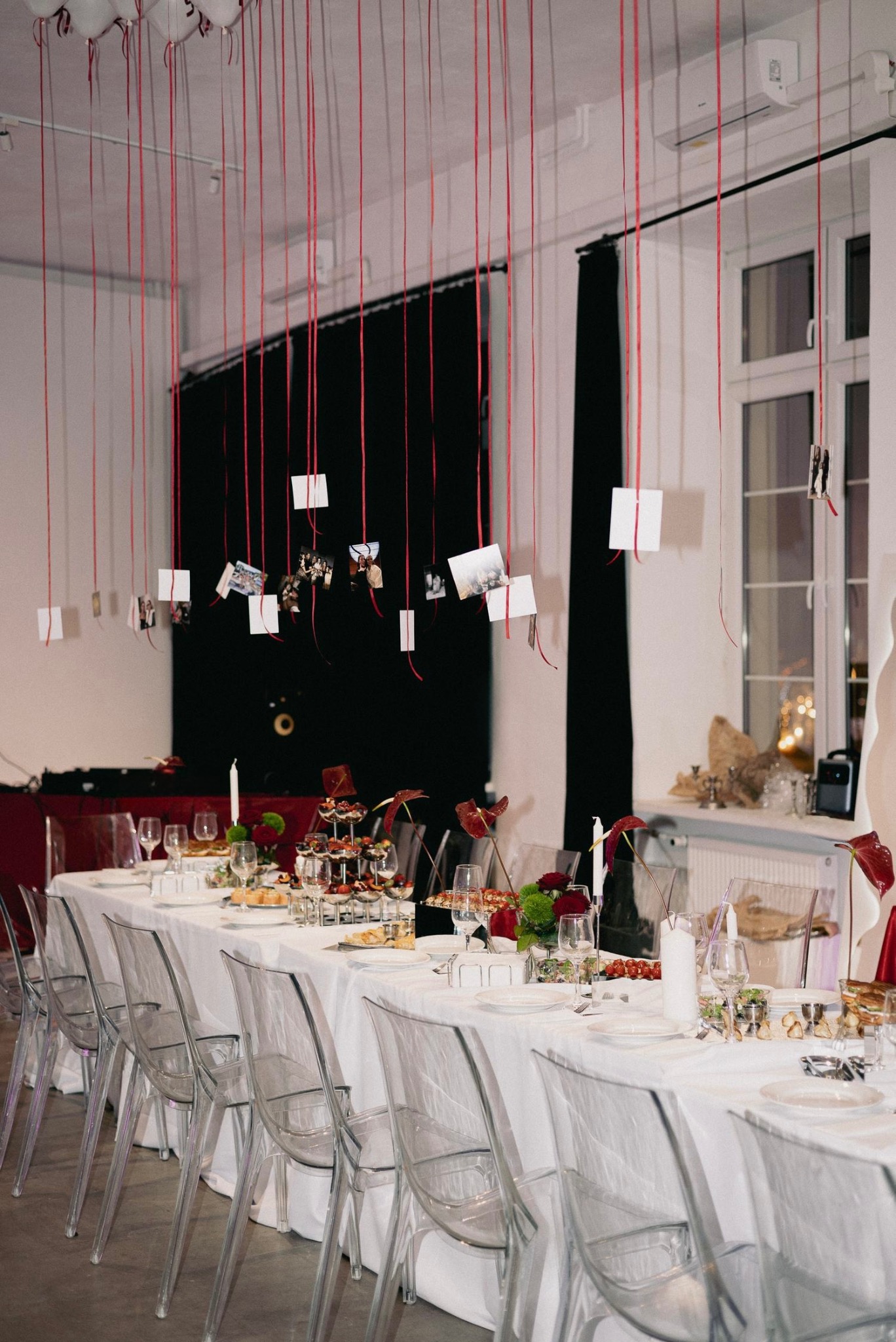 Uroczysty stół z białym obrusem, przezroczystymi krzesłami i dekoracjami z czerwonych wstążek z zawieszonymi zdjęciami. Elegancka aranżacja na imprezę.