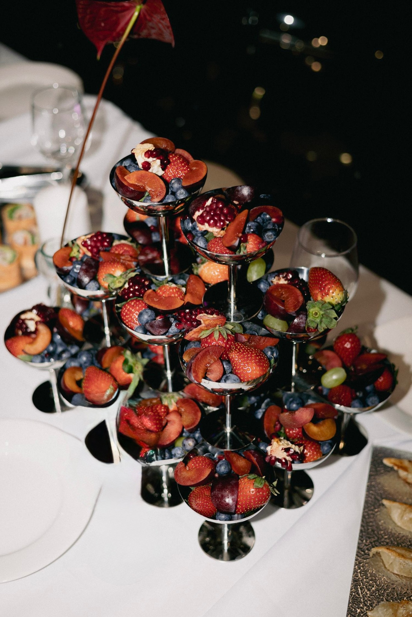 Elegancka, srebrna etażerka z owocami: truskawki, śliwki, borówki, granat. Dekoracja stołu na przyjęciu, widoczne kieliszki i przekąski w tle.