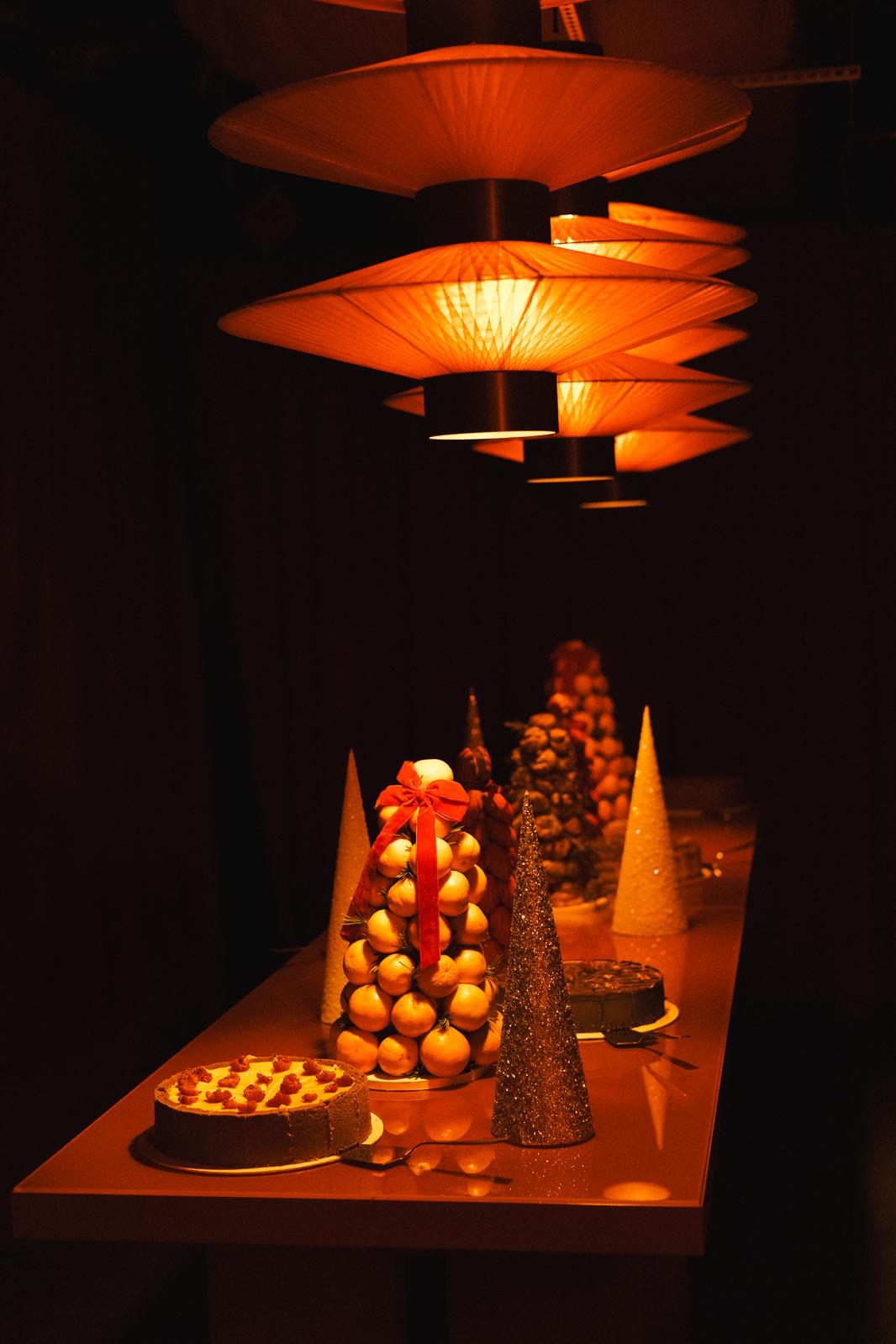 Świąteczny stół z tortami i piramidą z pomarańczy, udekorowany czerwoną wstążką, pod ciepłym światłem lamp. Dekoracje w kształcie choinek.