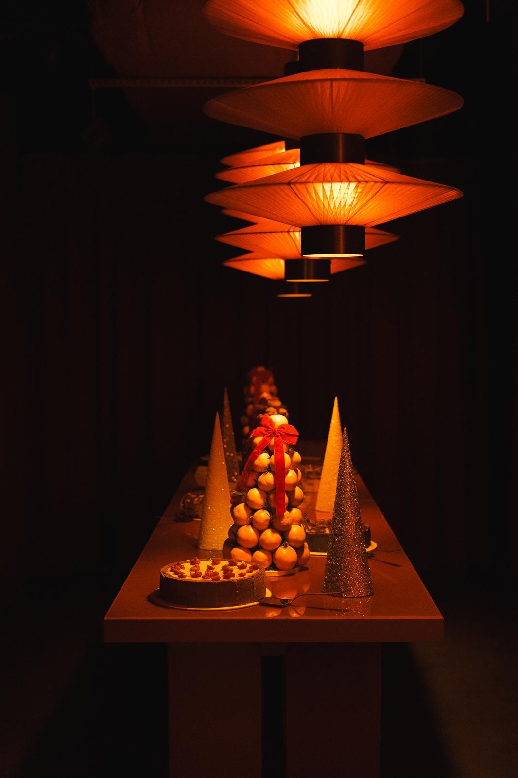 Świąteczny stół z tortem, piramidą z mandarynek z czerwoną kokardą i złotymi stożkami dekoracyjnymi pod ciepłym światłem lamp. Elegancki catering na imprezę.