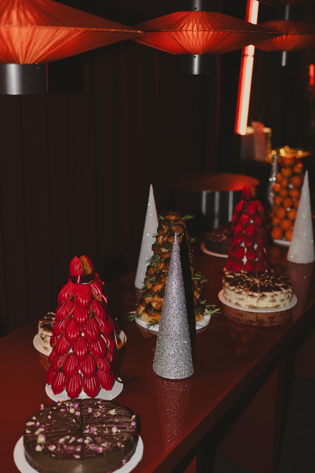 Dekoracyjny stół z ciastami i deserami w kształcie choinek, oświetlony ciepłym światłem, tworzący świąteczny nastrój. Ciemne tło podkreśla słodkości.