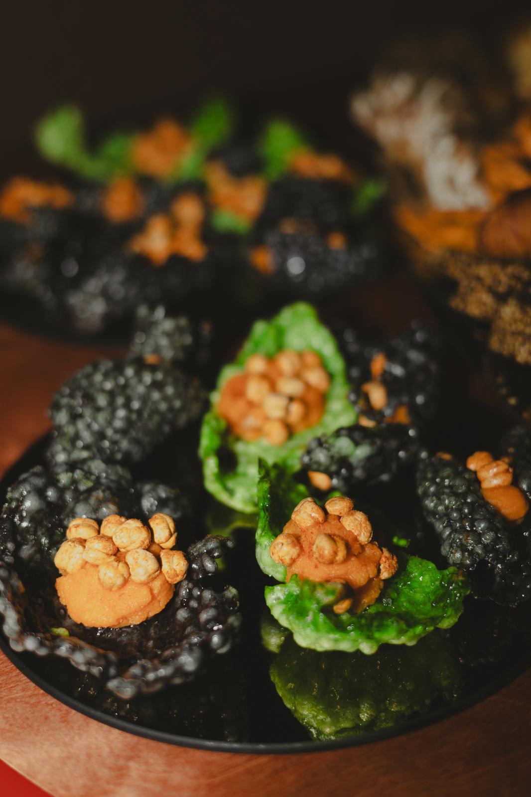 Dekoracyjne przekąski na czarnym talerzu: czarne chipsy z kawiorem i zielone z kremem i orzechami, zbliżenie, ciemne tło, elegancki catering.