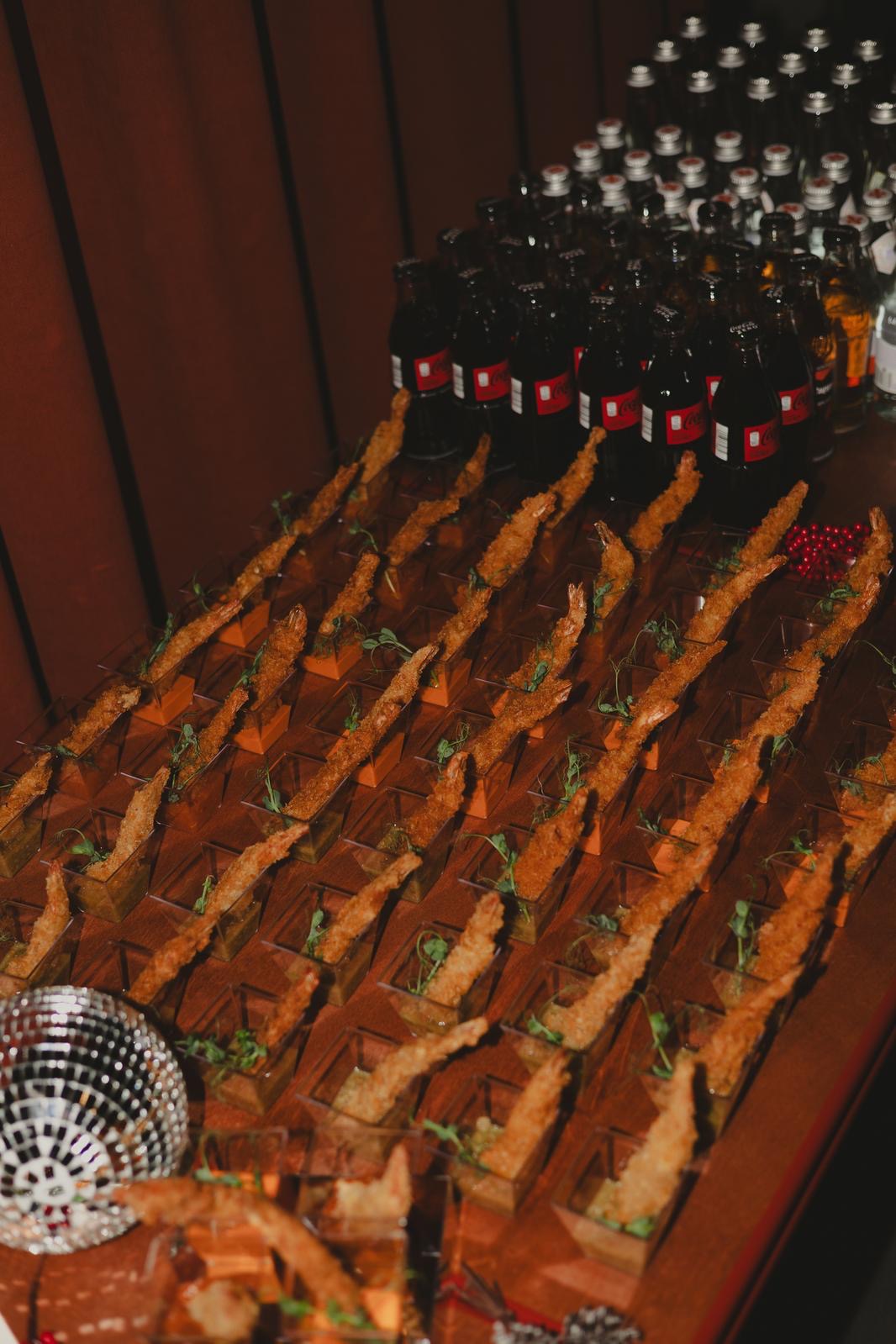 Elegancki catering: rzędy smażonych krewetek w małych naczyniach i butelki napojów na drewnianym stole, obok kula dyskotekowa. Przyciemnione światło.