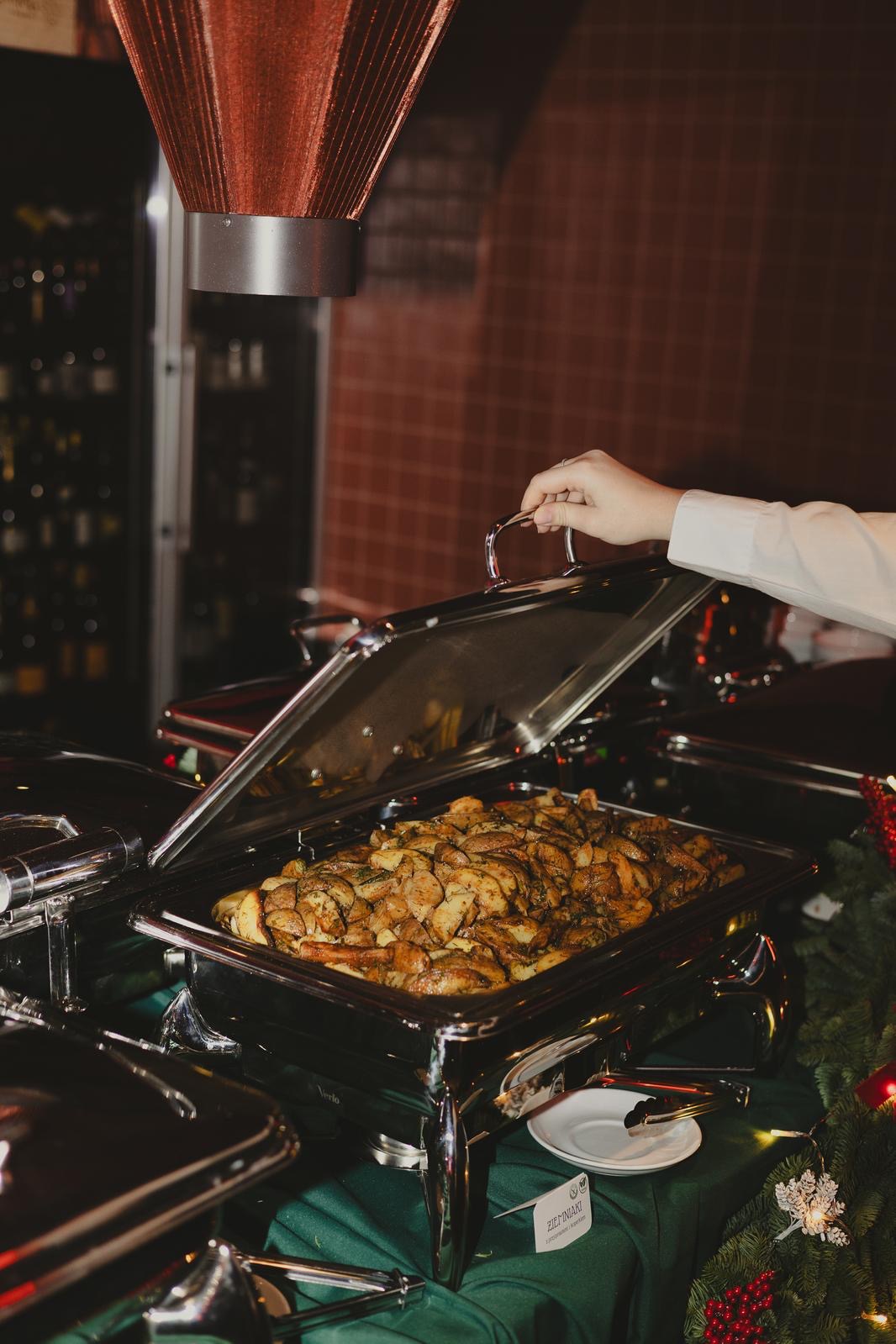 Podgrzewacz bufetowy ze złotymi ziemniakami, otwarty przez dłoń w białej koszuli. Dekoracja świąteczna, zielony obrus. Elegancki catering.