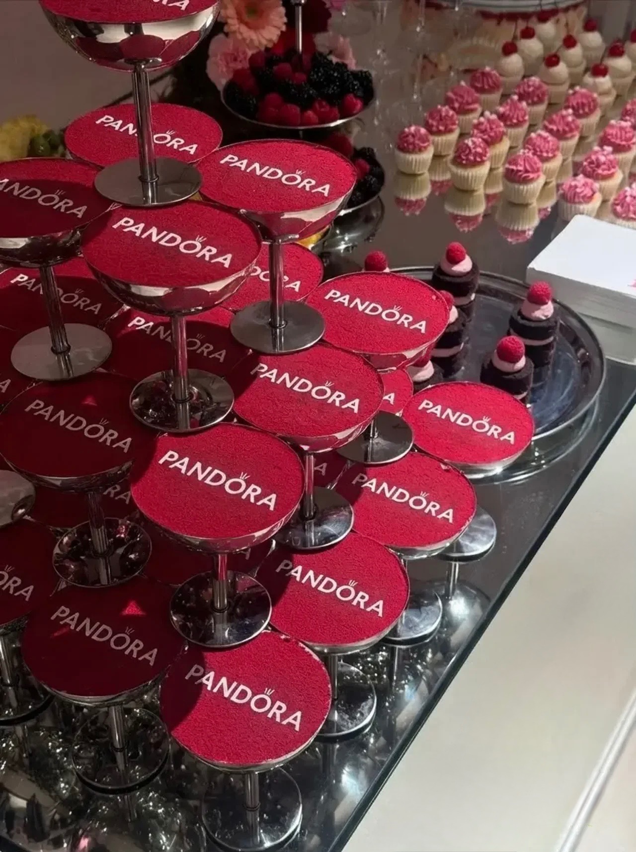 Piramida z kieliszków z czerwonym proszkiem i logo Pandora, obok babeczki i owoce na srebrnej tacy. Elegancki catering na imprezę firmową.
