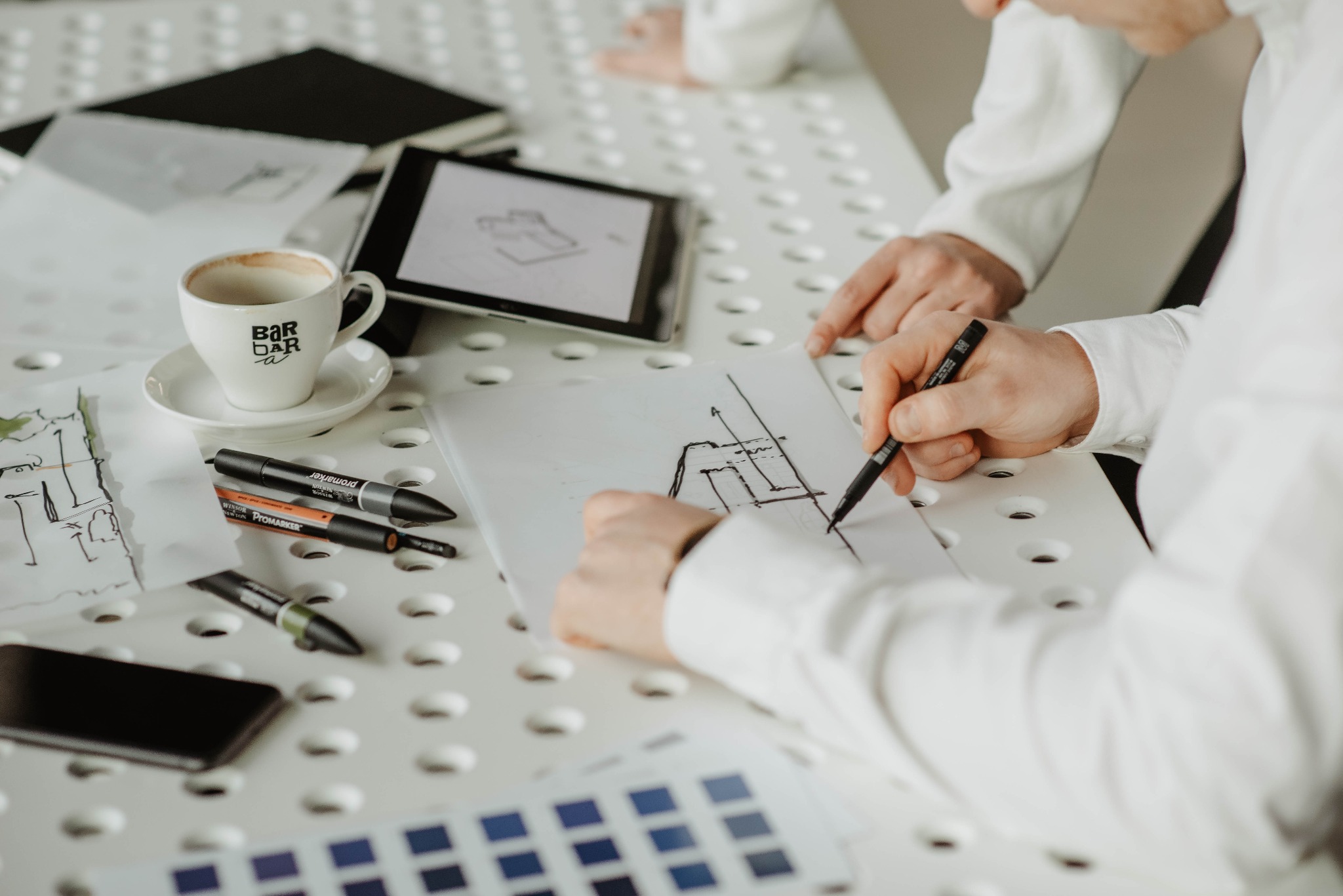 Architekt w białej koszuli rysuje plan budynku na białej kartce, obok tablet z widocznym szkicem, filiżanka kawy i markery na perforowanym stole.