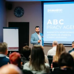 Prelegent w błękitnej marynarce prezentuje slajd 'ABC Pracy Agenta' na warsztatach mentoringowych o leczeniu za granicą, przed zgromadzonym audytorium. Widoczny zegar na ścianie i flipcharty...