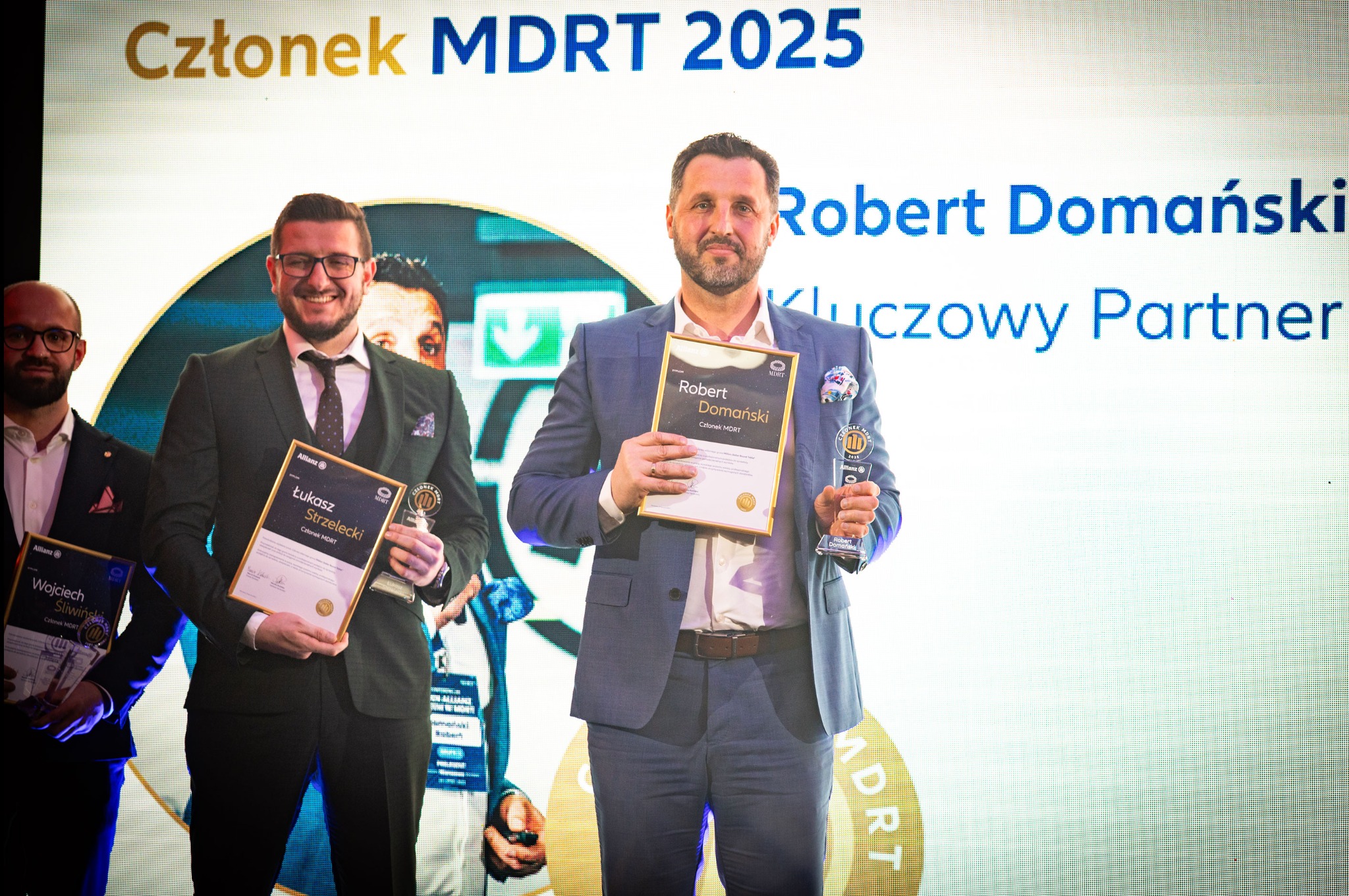 Trzej mężczyźni w garniturach pozują na tle ekranu z napisem 'Członek MDRT 2025', trzymając certyfikaty i statuetkę, wskazujące na osiągnięcia w branży finansowej.