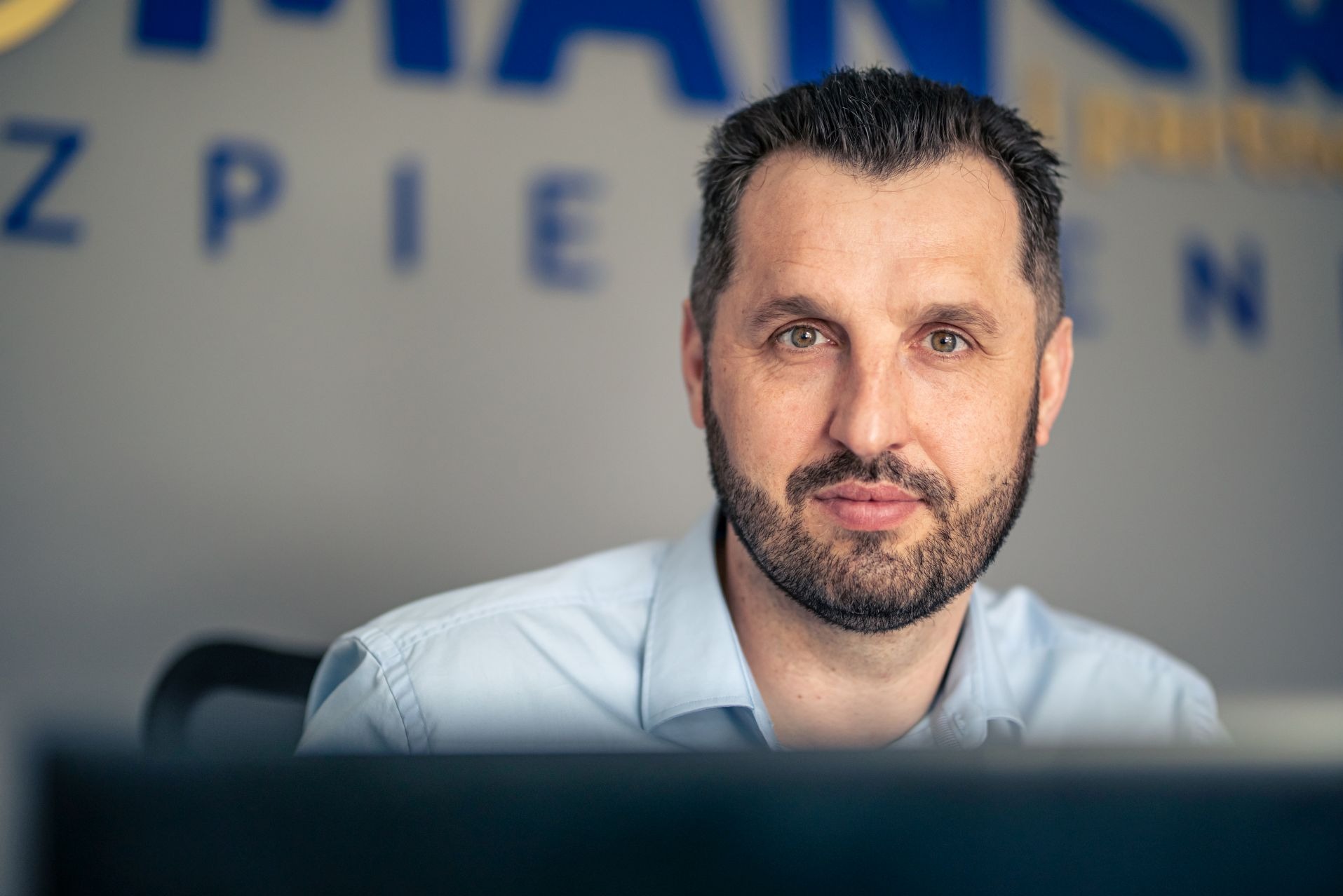 Portret mężczyzny z brodą w jasnoniebieskiej koszuli, spoglądającego w obiektyw na tle biurowym z rozmytym napisem.