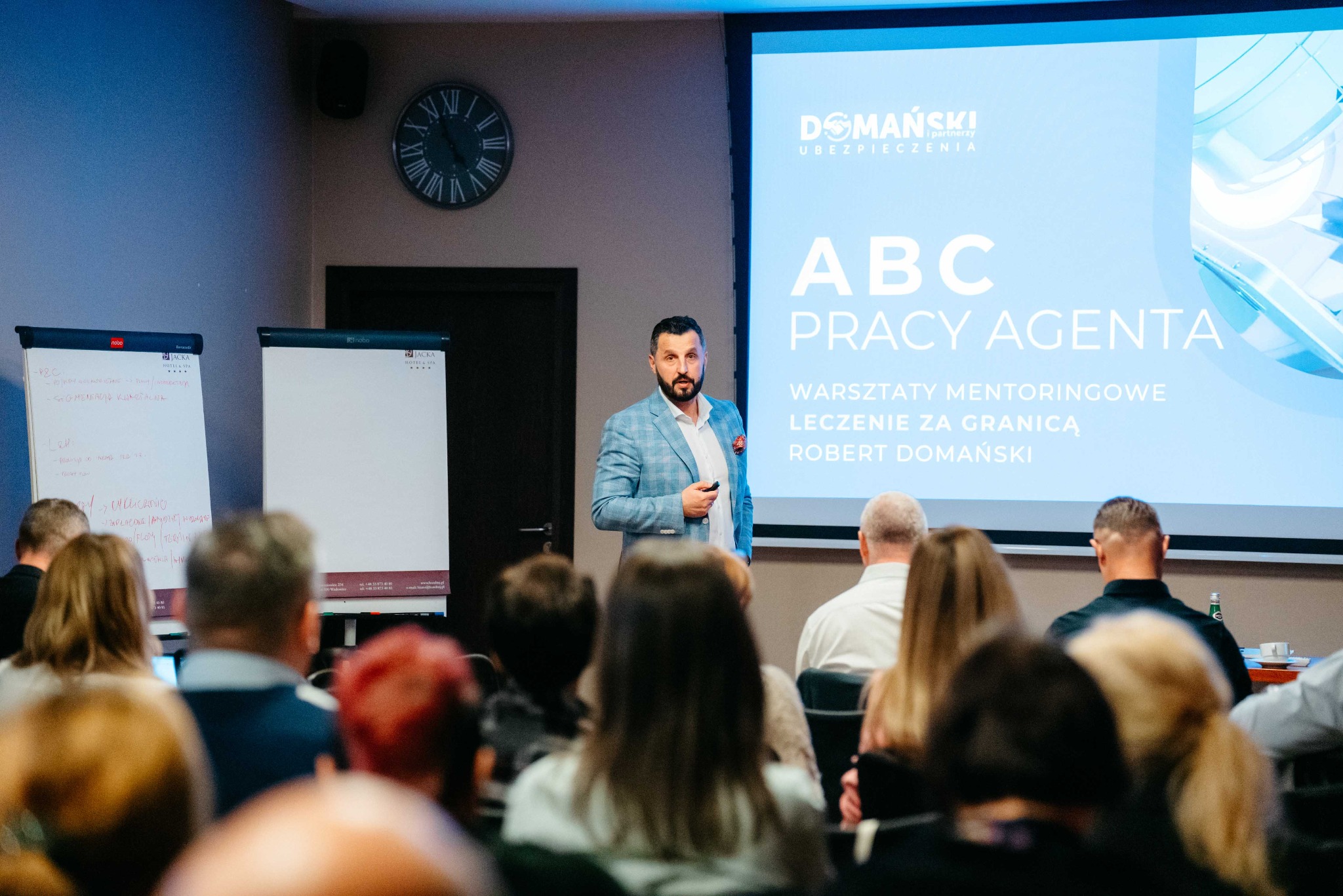 Prelegent w błękitnej marynarce prezentuje slajd 'ABC Pracy Agenta' na warsztatach mentoringowych o leczeniu za granicą, przed zgromadzonym audytorium. Widoczny zegar na ścianie i flipcharty...