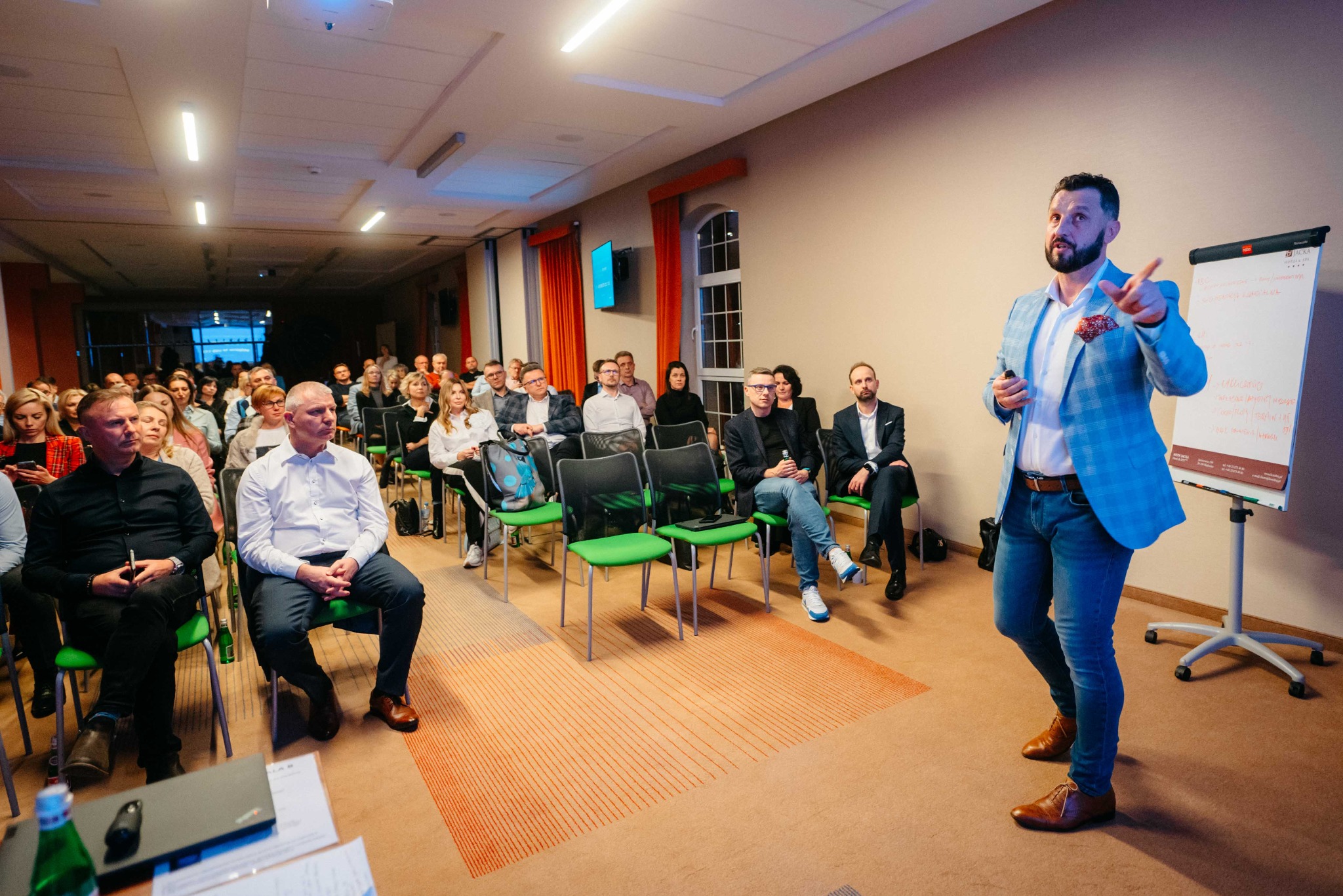 Prelegent w niebieskiej marynarce gestykuluje podczas prezentacji dla publiczności siedzącej na zielonych krzesłach w sali konferencyjnej; flipchart z notatkami po prawej.