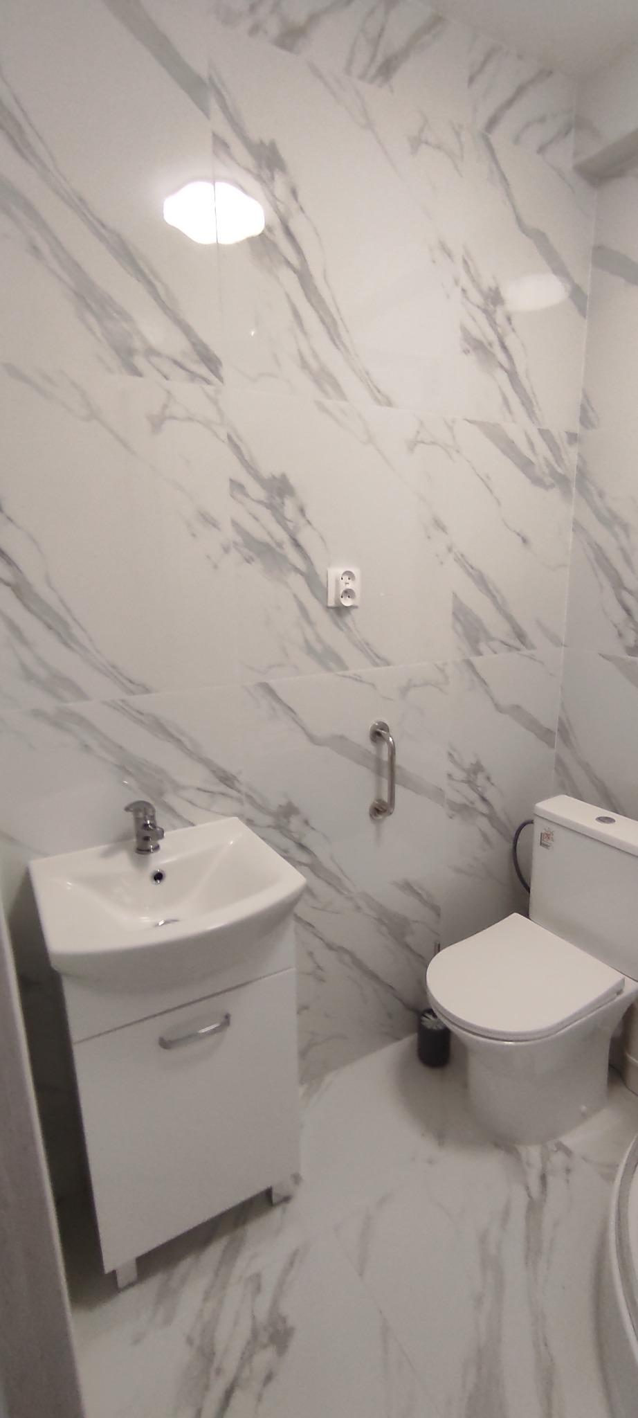 Biała łazienka z marmurowymi płytkami, umywalką z szafką, toaletą i uchwytem dla niepełnosprawnych. Nowoczesny design, czystość i funkcjonalność.