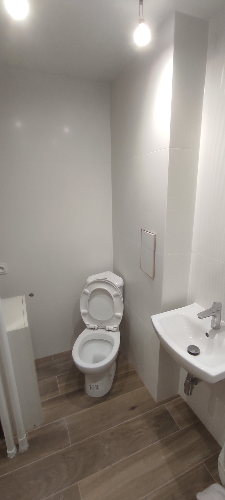 Jasna łazienka po remoncie: biała ceramika (toaleta z otwartą klapą, umywalka), drewnopodobne płytki podłogowe, minimalistyczne wykończenie.