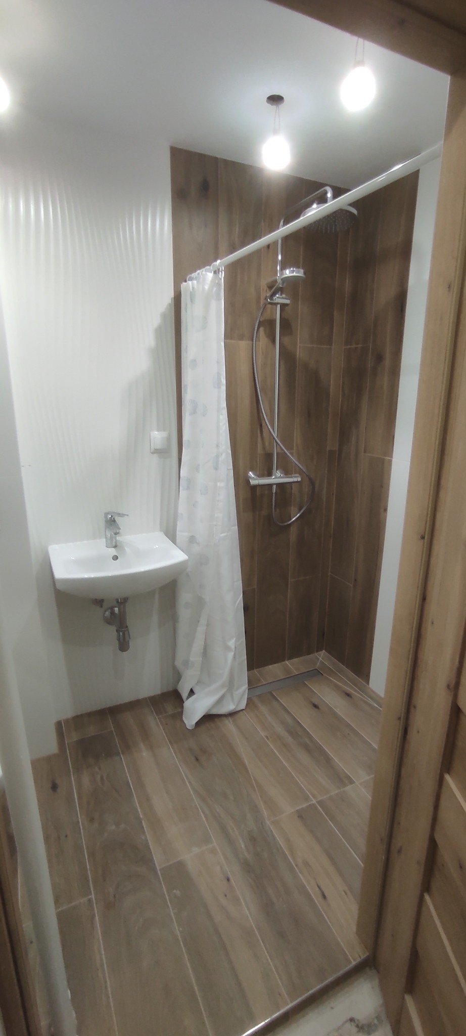 Nowoczesna łazienka z kabiną prysznicową typu walk-in, wykończona płytkami imitującymi drewno, z białą umywalką i zasłoną prysznicową w jasnym odcieniu.