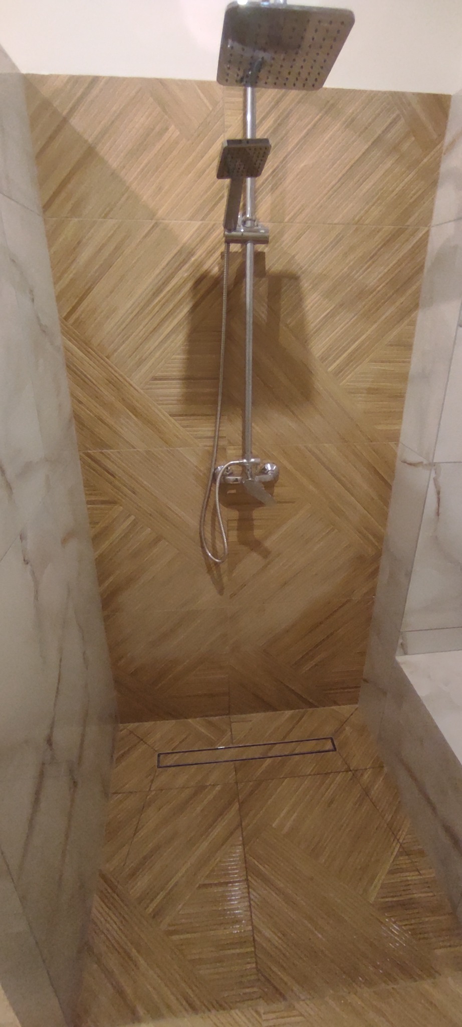 Kabina prysznicowa z drewnopodobnymi płytkami ułożonymi w jodełkę, odpływ liniowy i chromowana armatura. Marmurowe wykończenie ścian.