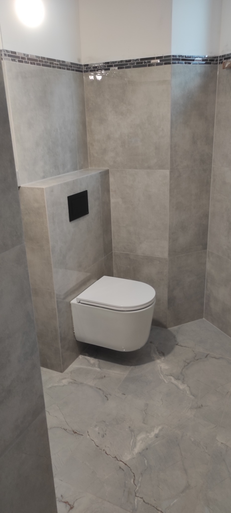 Wykończona łazienka w szarościach z podwieszaną toaletą i mozaiką na górze płytek. Płytki imitujące marmur na podłodze i ścianach. Minimalistyczny styl.