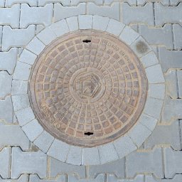 Firma usługowo-budowlana NIKITA Paulina Walczyńska - Szara kostka brukowa typu 'I' z okrągłą studzienką kanalizacyjną otoczoną mniejszą kostką. Widoczny fragment buta w lewym dolnym rogu.