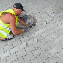 Firma usługowo-budowlana NIKITA Paulina Walczyńska - Pracownik w odblaskowej kamizelce i czapce układa szarą kostkę na chodniku, używając gumowego młotka w pobliżu studzienki kanalizacyjnej.