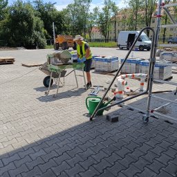 Firma usługowo-budowlana NIKITA Paulina Walczyńska - Pracownik w kamizelce odblaskowej i kapeluszu przy maszynie do cięcia kostki brukowej na placu budowy, w tle zaparkowany wywrotka i budynek mieszkalny.