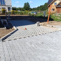 Firma usługowo-budowlana NIKITA Paulina Walczyńska - Układanie szarej kostki brukowej typu 'puzzle' na posesji, widoczne fragmenty podbudowy z kruszywa i elementy obrzeży betonowych, w tle dom w stanie surowym i drewniany domek letniskowy.