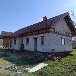 Firma usługowo-budowlana NIKITA Paulina Walczyńska - Surowy stan domu jednorodzinnego w trakcie budowy: ściany z białych bloczków, czerwona dachówka, widoczne okna i rusztowanie, trawa na pierwszym planie.