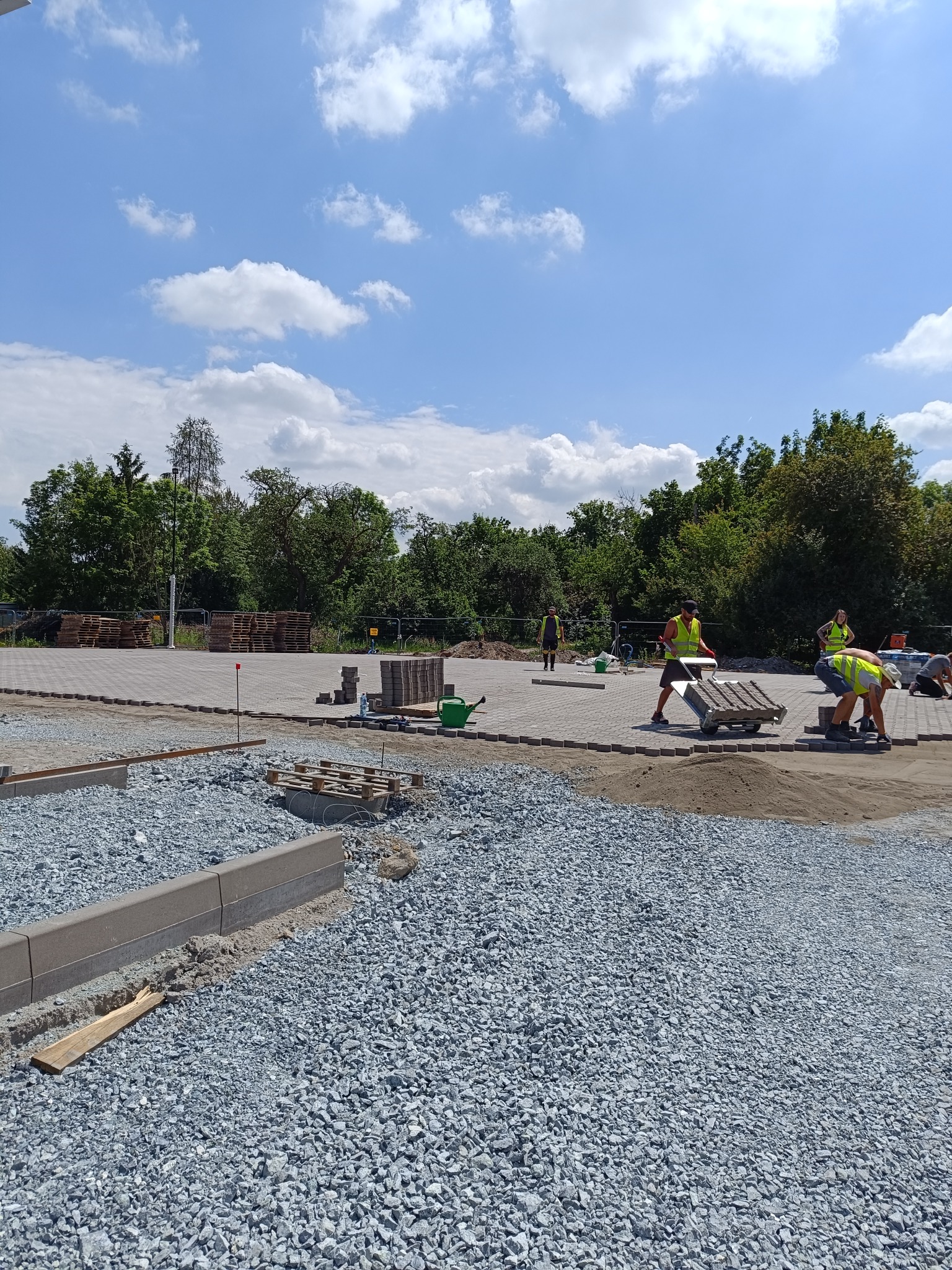 Ekipa w kamizelkach odblaskowych układa szarą kostkę na placu; widoczny stos kostki, taczka z kostką i podsypka z szarego grysu.