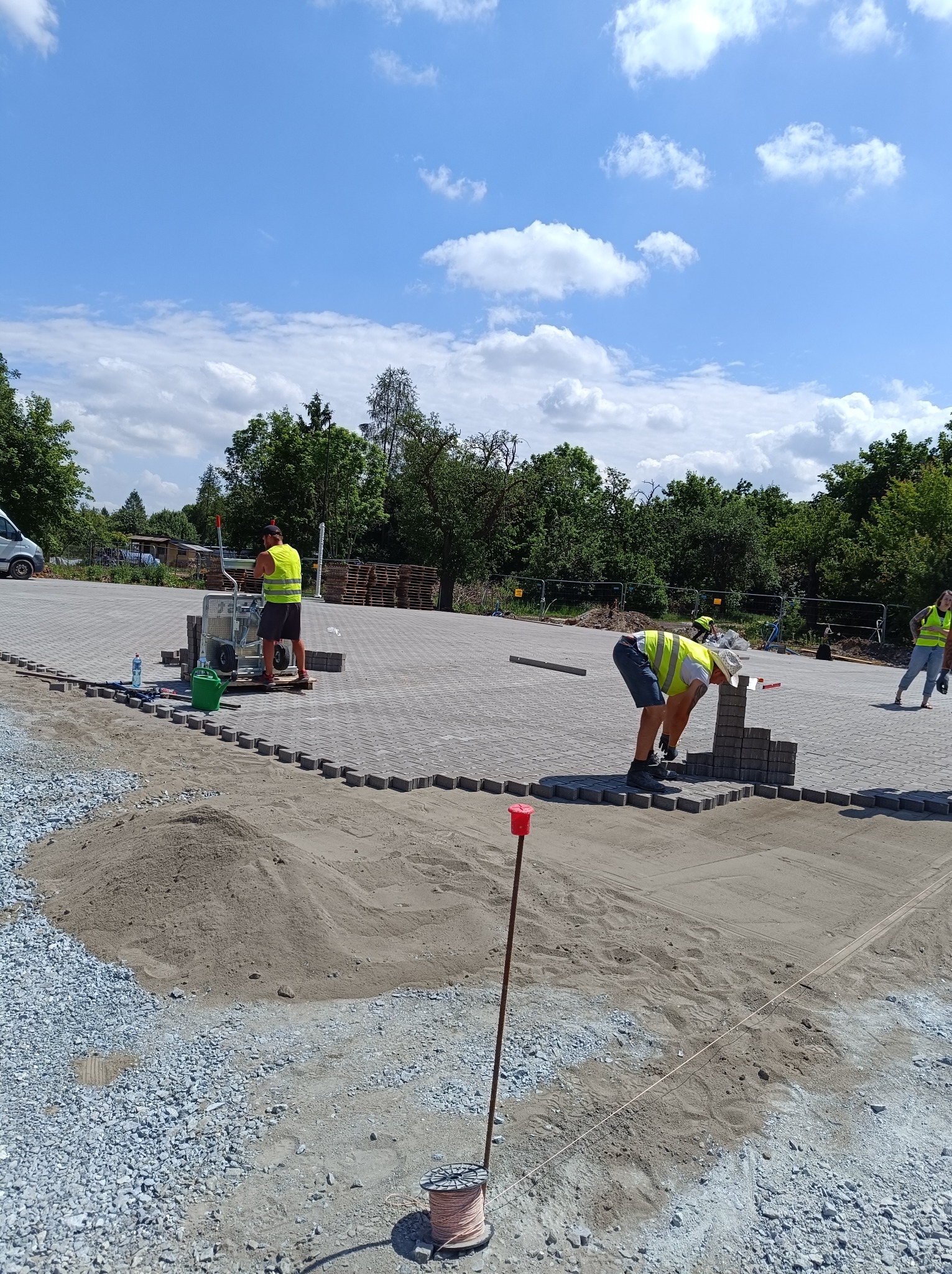Pracownicy w kamizelkach odblaskowych układają szarą kostkę na przygotowanym podłożu. W tle widoczny sprzęt budowlany, palety i zieleń drzew.