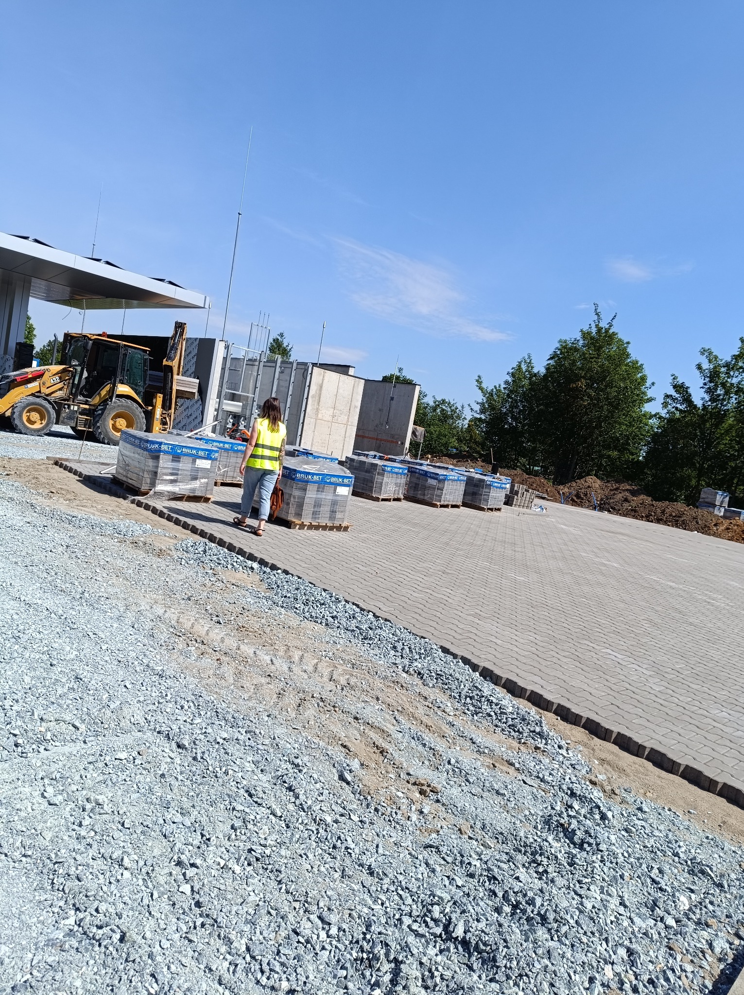 Układanie kostki brukowej: kobieta w kamizelce odblaskowej przechodzi obok palet z kostką Bruk-Bet, w tle żółta ładowarka i częściowo ułożona nawierzchnia z kostki.