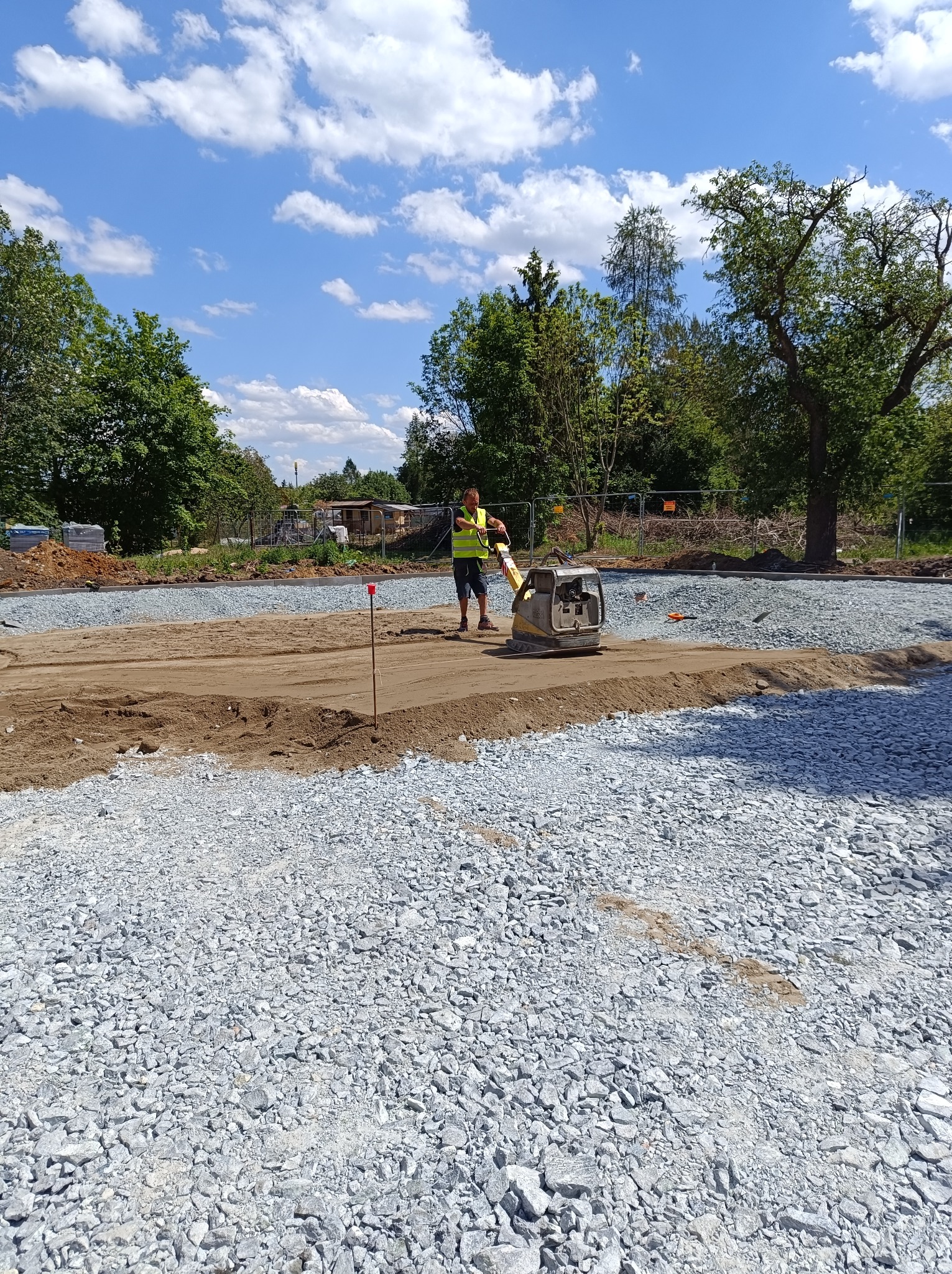 Pracownik w żółtej kamizelce ubija piasek zagęszczarką na placu budowy, gdzie w tle widoczne są ułożone warstwy kruszywa i drzewa.