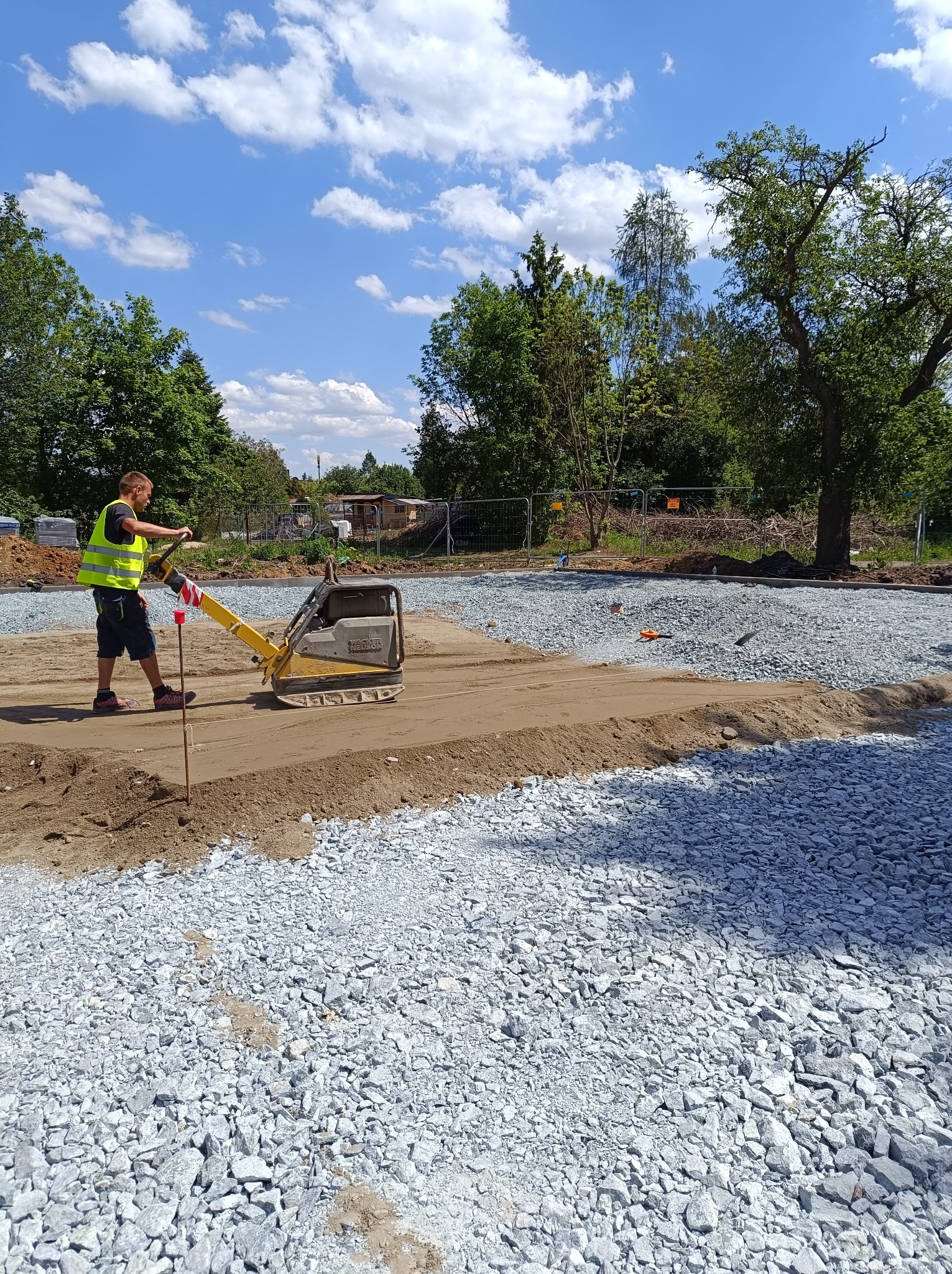Pracownik w kamizelce odblaskowej zagęszcza piasek na podbudowie z kruszywa, przygotowując teren pod nawierzchnię.
