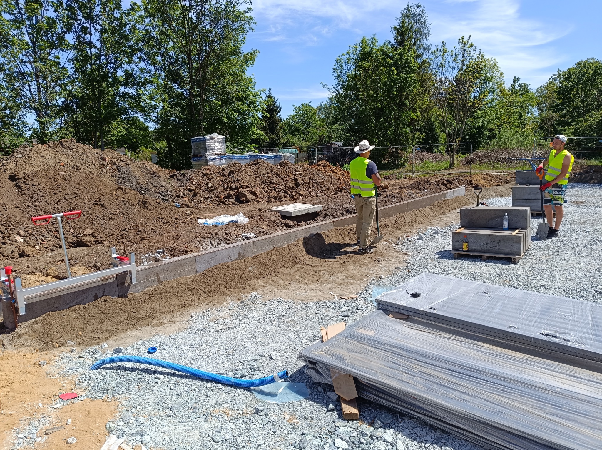 Dwaj pracownicy w kamizelkach odblaskowych przygotowują teren pod budowę chodnika z betonowych płyt, widoczne narzędzia, piasek, ziemia i stos płyt na palecie.