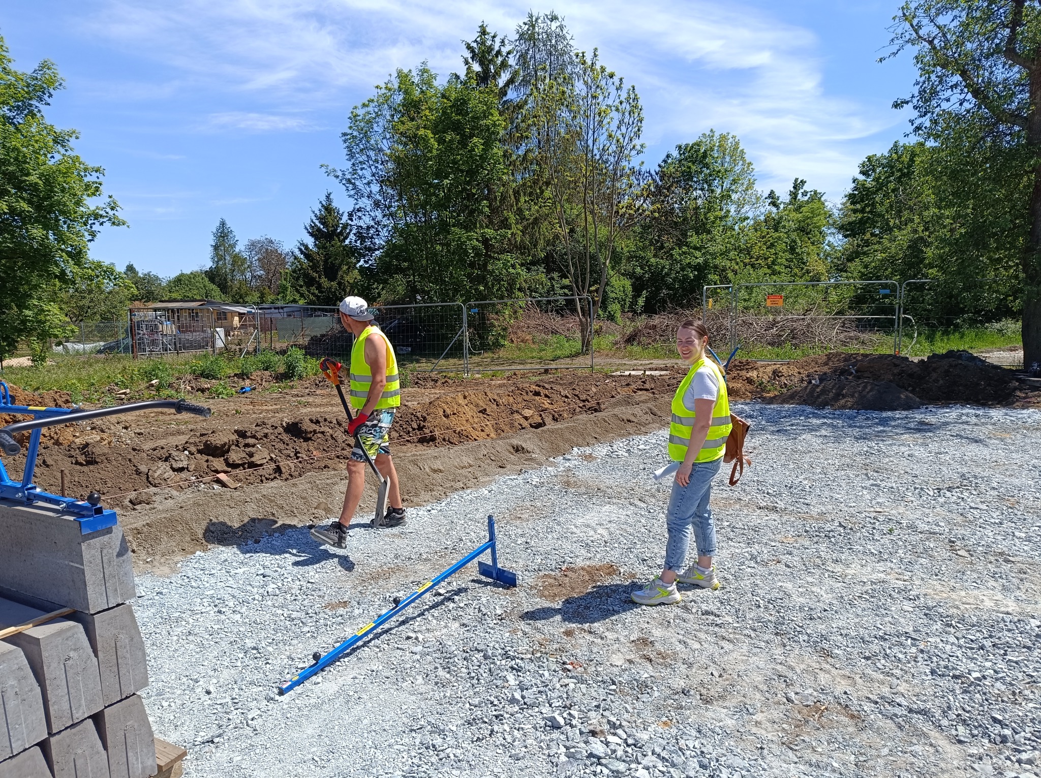 Przygotowanie podłoża pod kostkę: dwójka pracowników w kamizelkach odblaskowych na placu budowy z podsypką żwirową, w tle ułożone obrzeża i zielone drzewa.