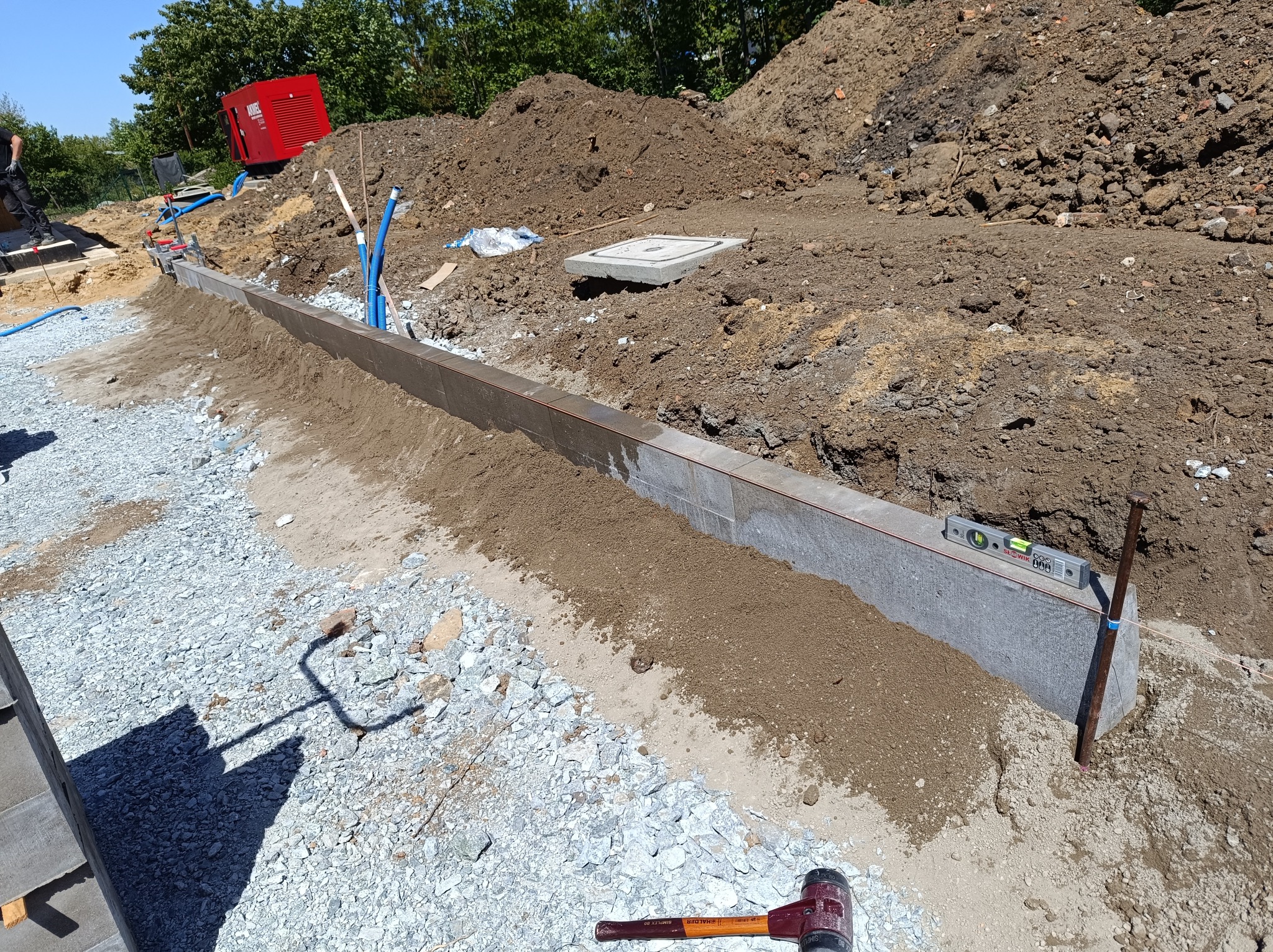 Przygotowanie podłoża pod krawężnik z użyciem poziomicy, widoczne ubite kruszywo, wbite szpilki, oraz gumowy młotek Halder.