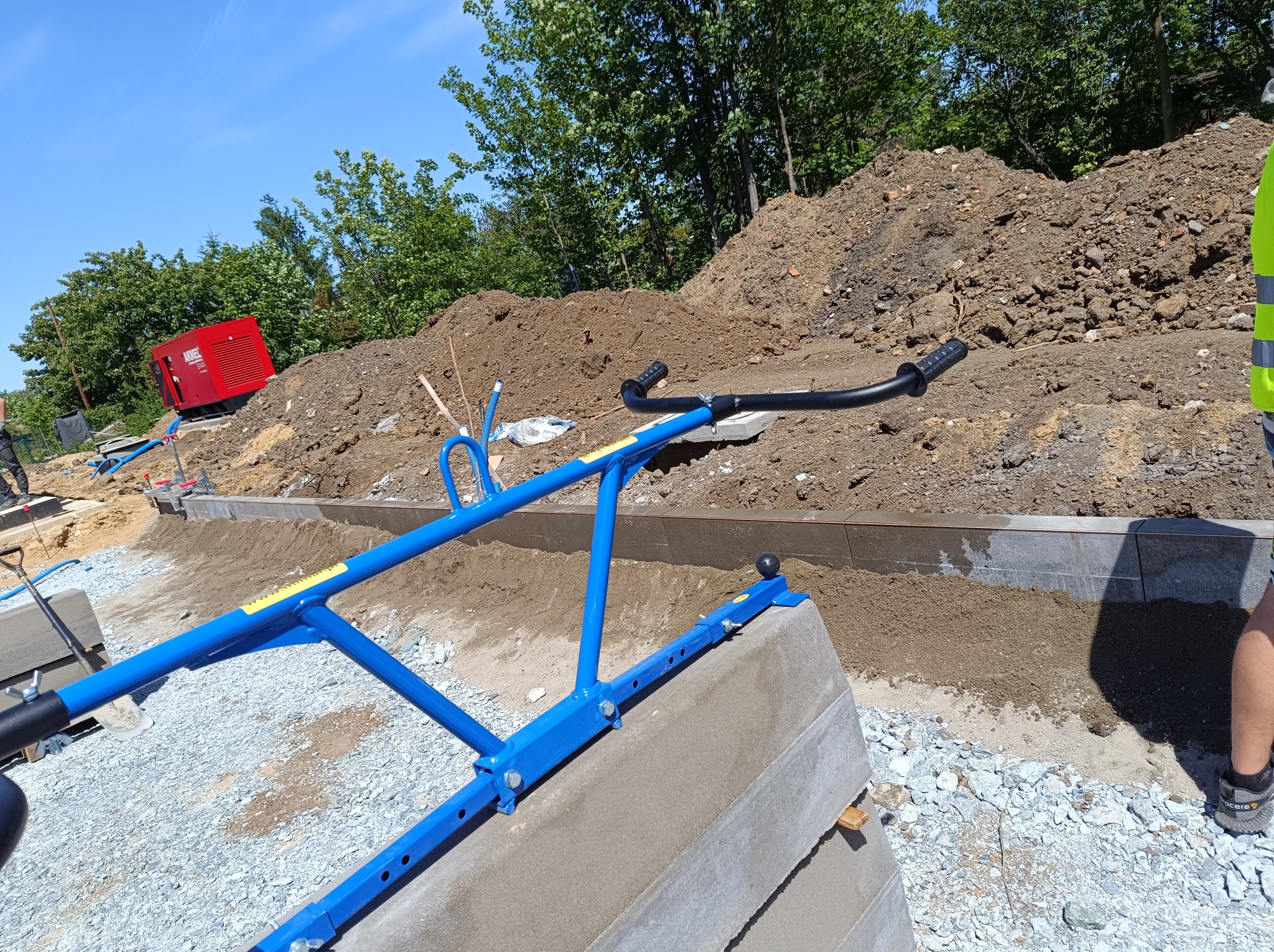 Niebieskie narzędzie do wyrównywania podłoża pod kostkę brukową, widoczny stos krawężników, przygotowany teren i hałda ziemi w tle.