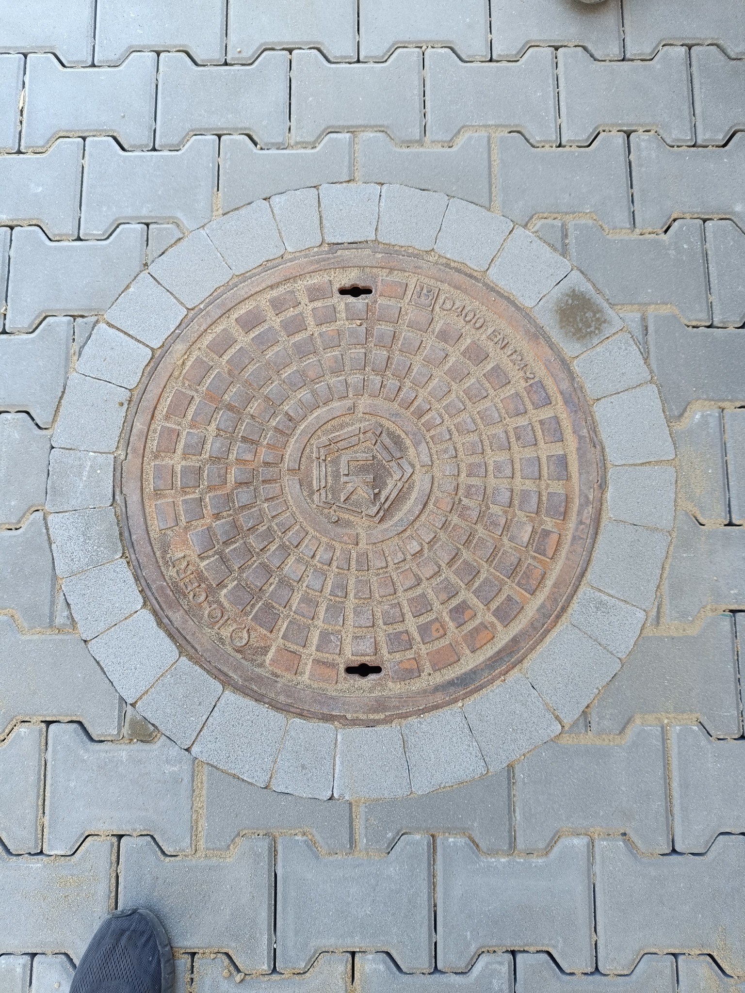 Szara kostka brukowa typu 'I' z okrągłą studzienką kanalizacyjną otoczoną mniejszą kostką. Widoczny fragment buta w lewym dolnym rogu.