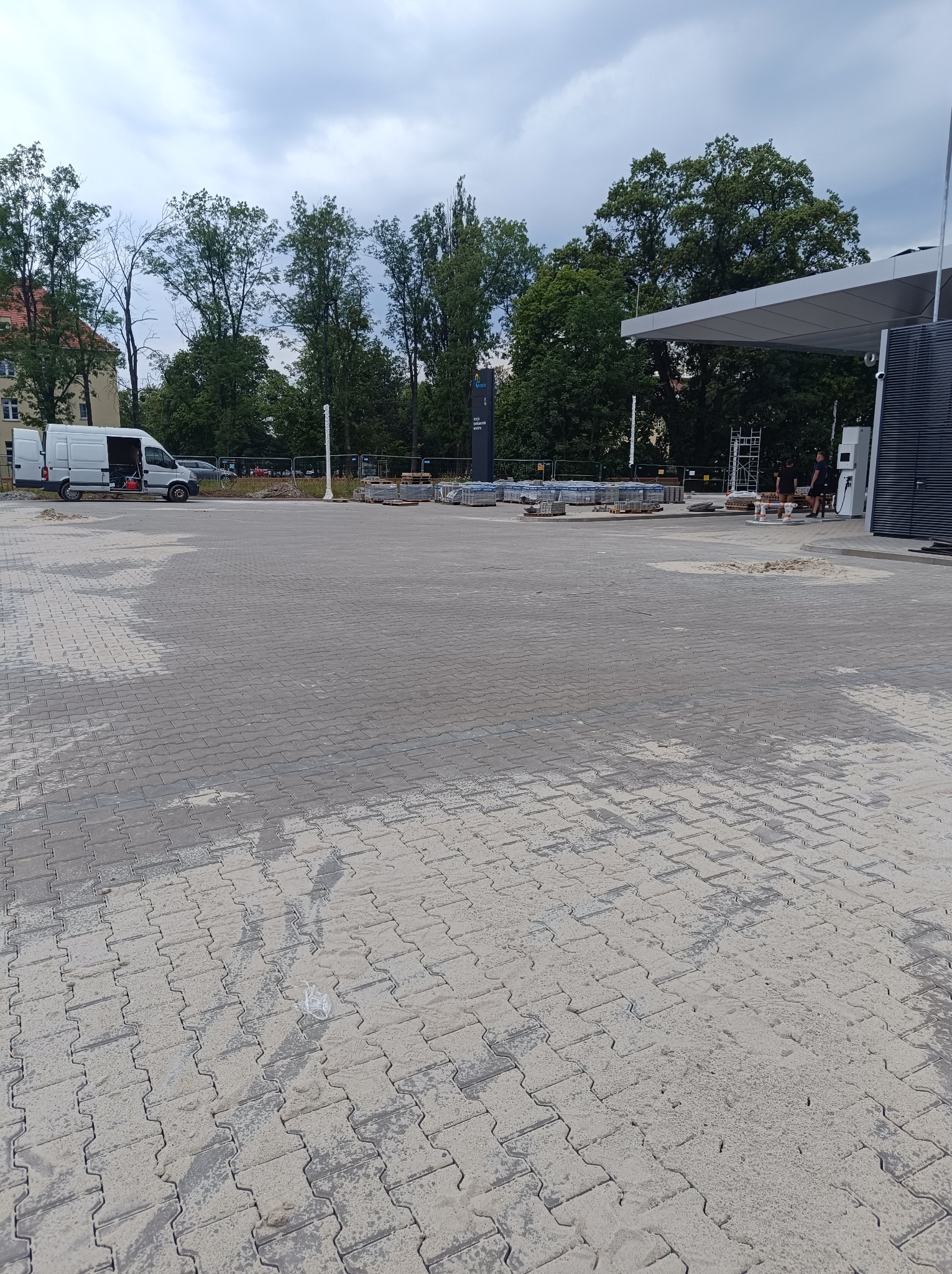 Szara kostka brukowa na placu budowy, w tle zaparkowany biały van i drzewa, widoczne ślady piasku i wody na powierzchni kostki.