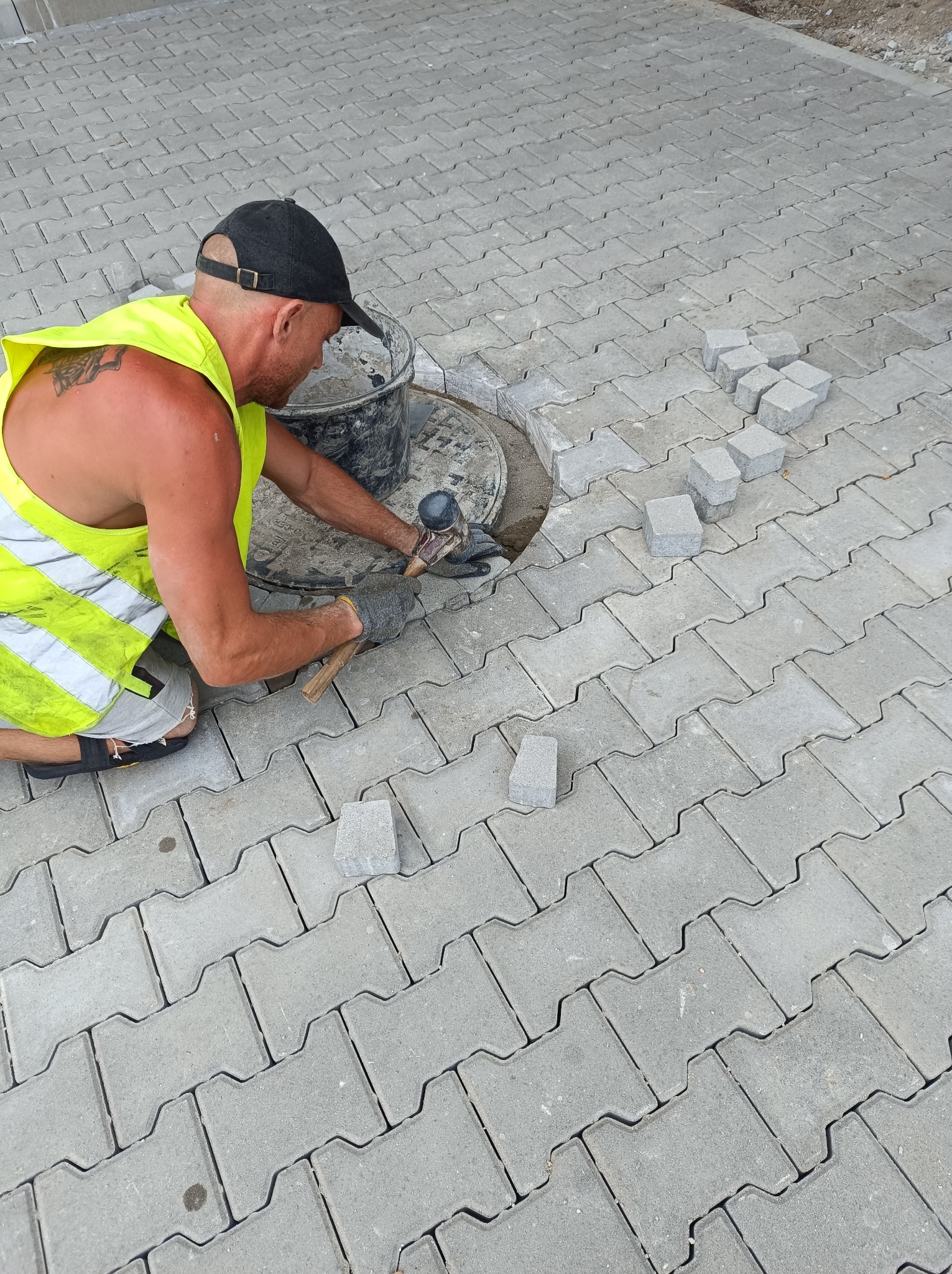 Pracownik w odblaskowej kamizelce i czapce układa szarą kostkę na chodniku, używając gumowego młotka w pobliżu studzienki kanalizacyjnej.