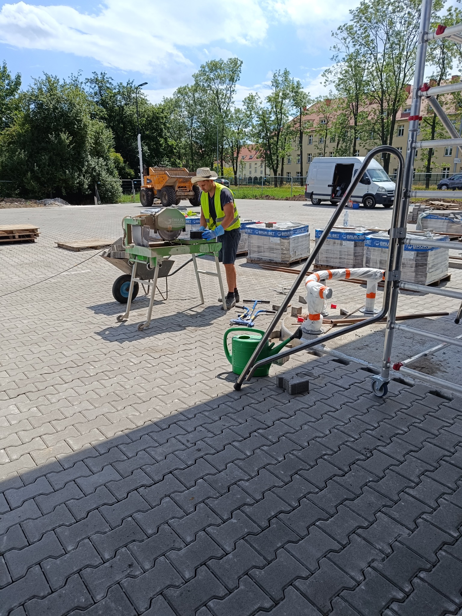 Pracownik w kamizelce odblaskowej i kapeluszu przy maszynie do cięcia kostki brukowej na placu budowy, w tle zaparkowany wywrotka i budynek mieszkalny.