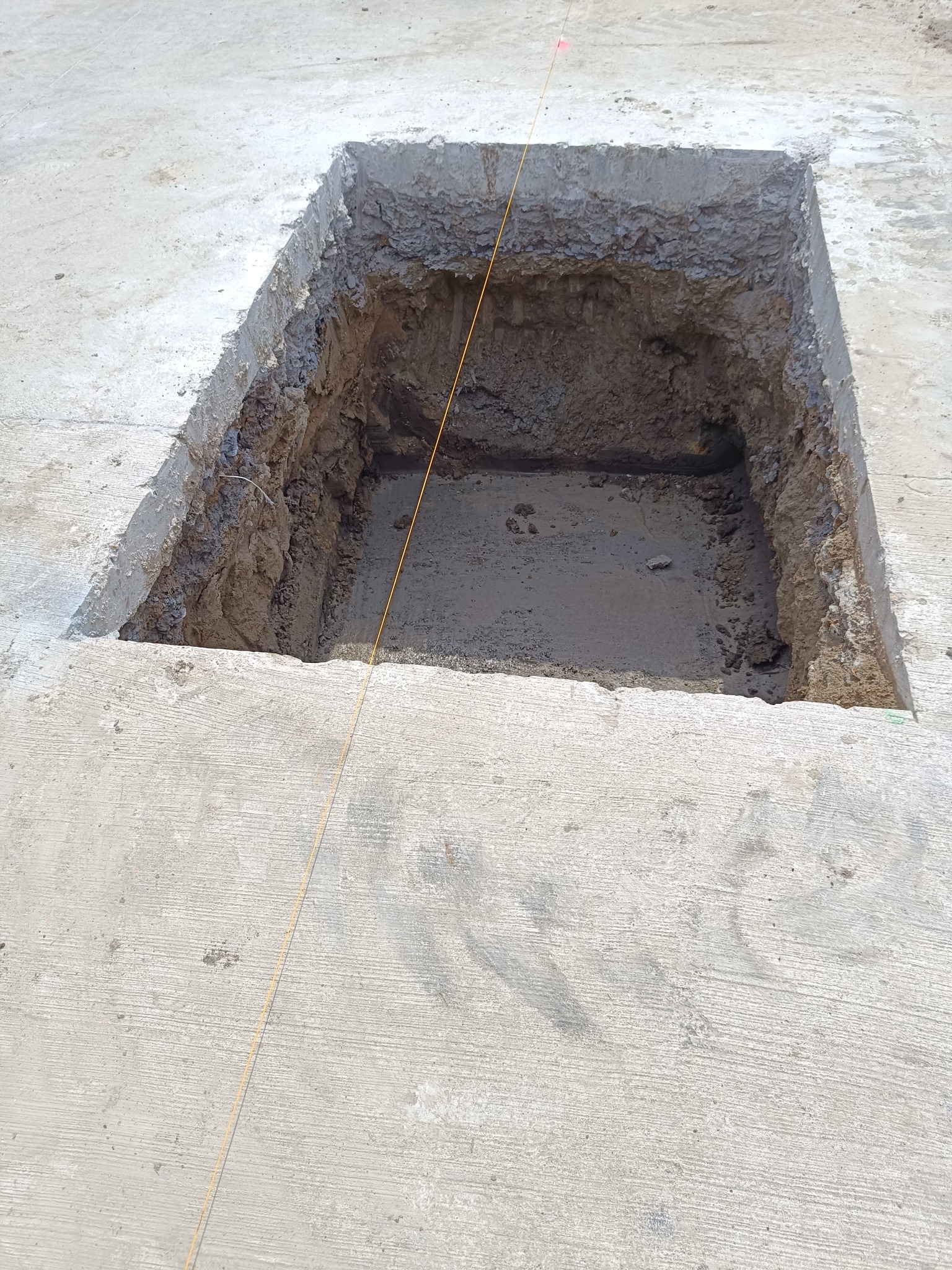 Kwadratowy wykop w betonowej powierzchni, widoczna ziemia wewnątrz, pomarańczowa linka wyznaczająca granice.