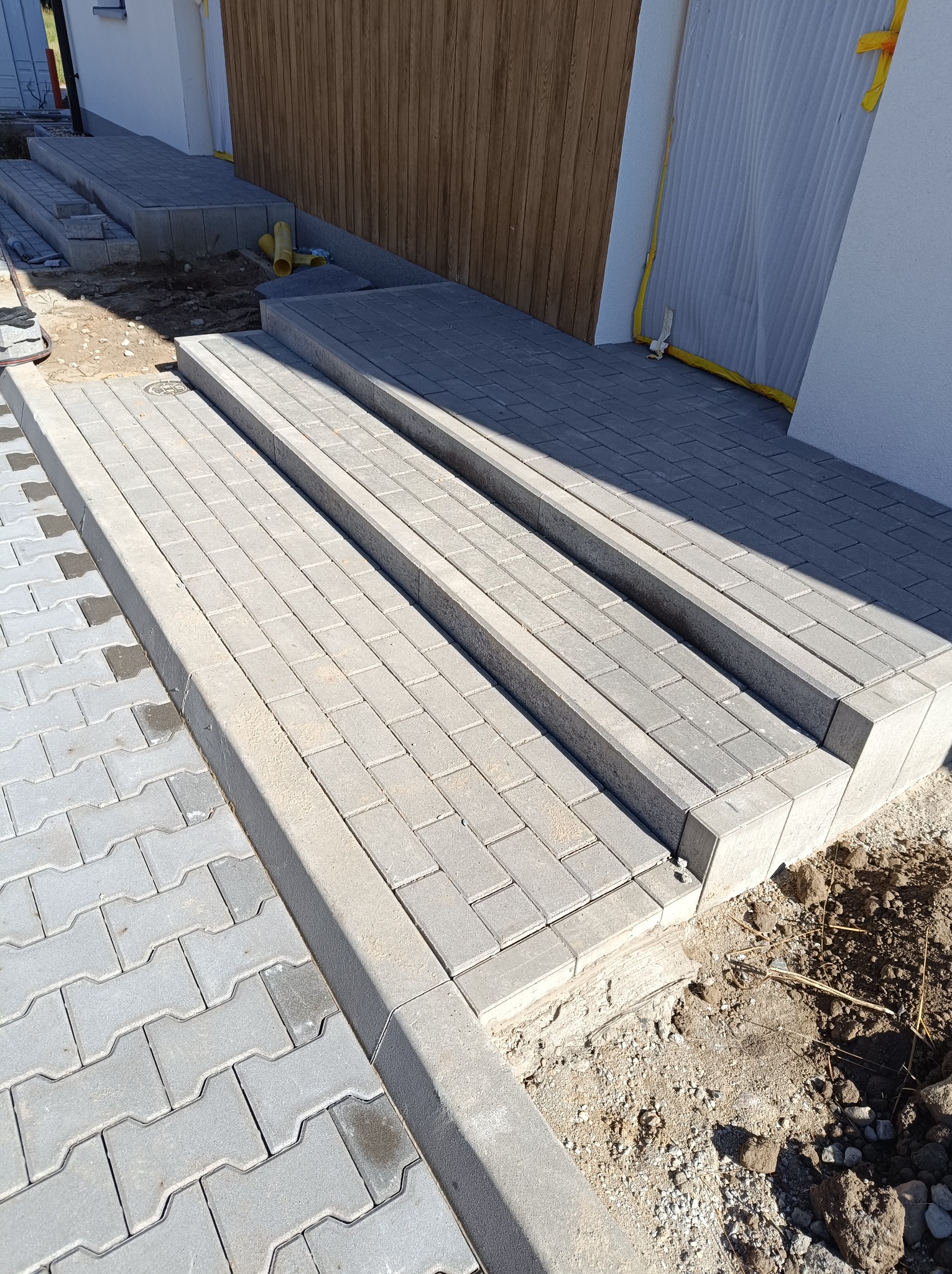 Schody wejściowe z szarej kostki brukowej prowadzące do budynku z drewnianą elewacją, widoczne niedokończone prace ziemne wokół schodów.