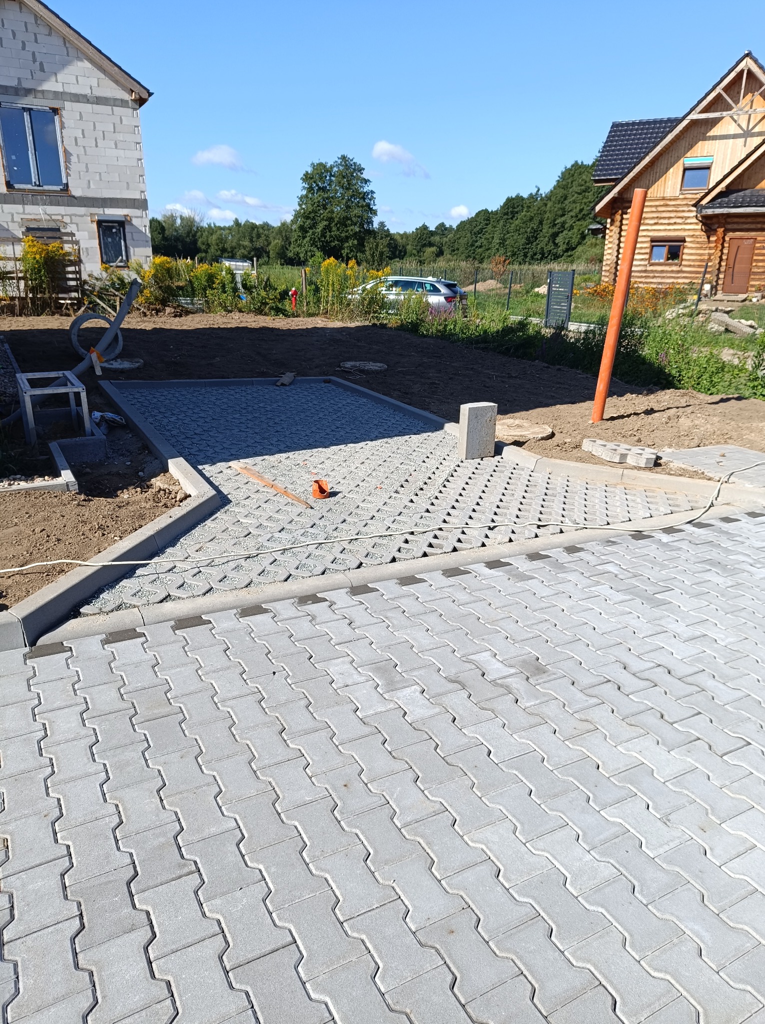 Układanie szarej kostki brukowej typu 'puzzle' na posesji, widoczne fragmenty podbudowy z kruszywa i elementy obrzeży betonowych, w tle dom w stanie surowym i drewniany domek letniskowy.