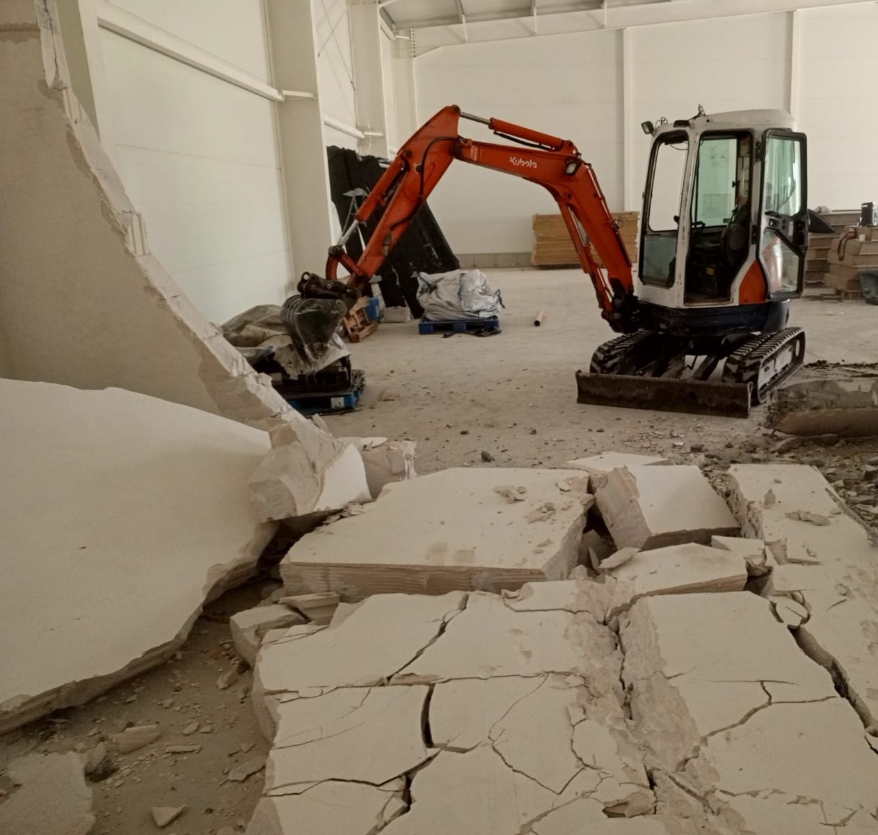 Pomarańczowa mini koparka Kubota kruszy betonową podłogę wewnątrz dużego, jasnego magazynu.