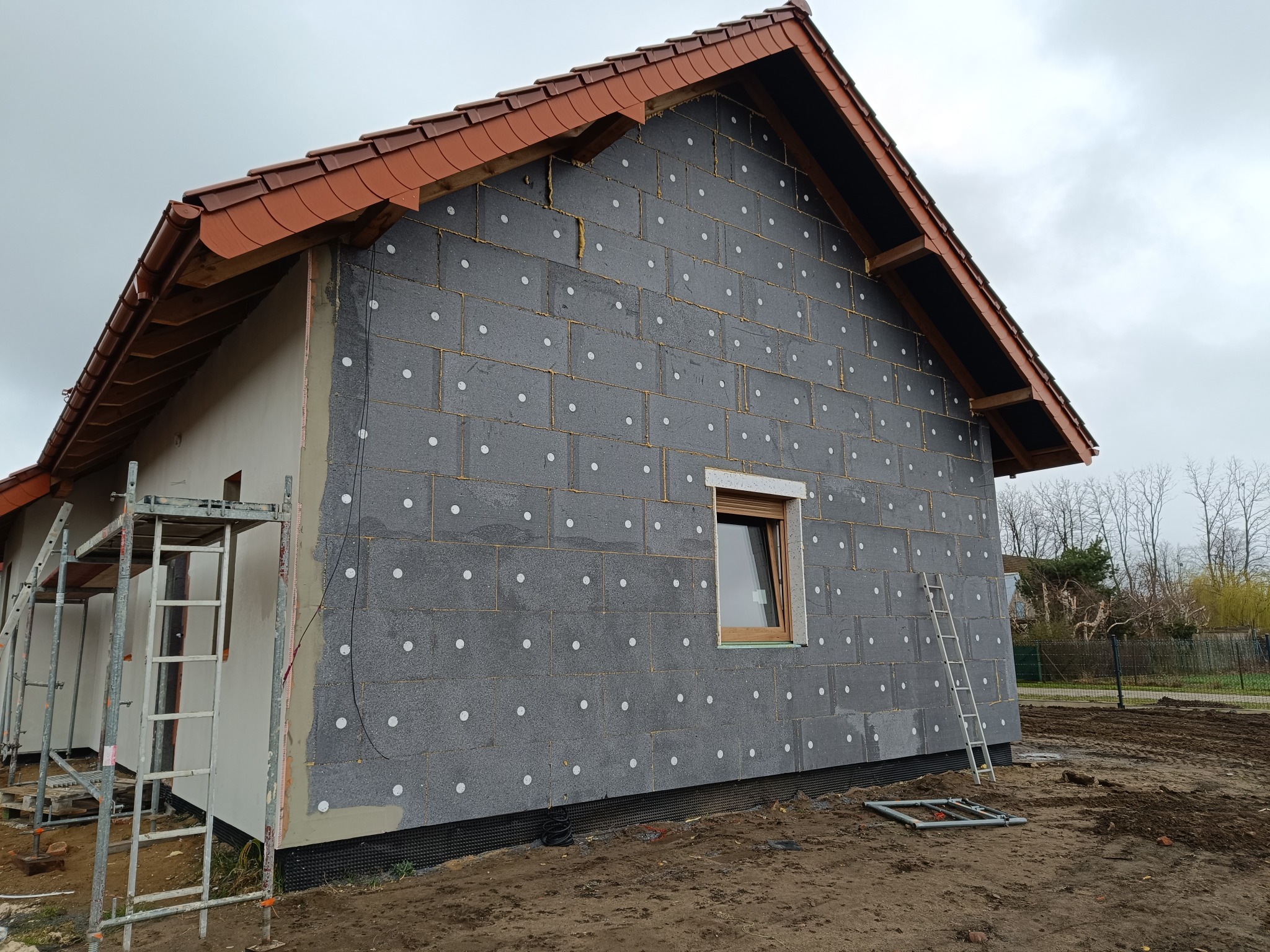 Dom jednorodzinny w trakcie ocieplania: szare bloki izolacyjne z białymi kołkami mocowane do ściany, widoczna drabina i rusztowanie, brązowy dach z dachówki ceramicznej, ziemisty teren wokół budynku.