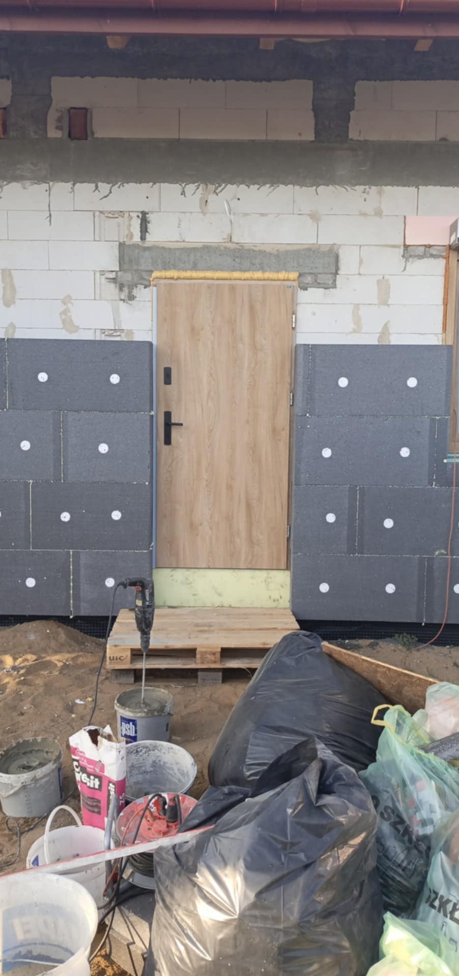 Budynek w trakcie ocieplania: szare płyty styropianowe z białymi kołkami częściowo przymocowane do ściany z białych bloczków, drewniane drzwi wejściowe zabezpieczone żółtą pianką montażową, paleta...