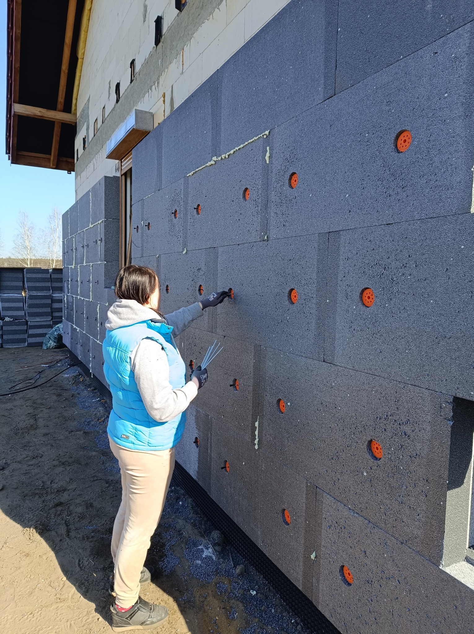 Pracownica w niebieskiej kamizelce montuje szare płyty izolacyjne na ścianie budynku, mocując je pomarańczowymi kołkami; widoczne zapasy płyt obok.