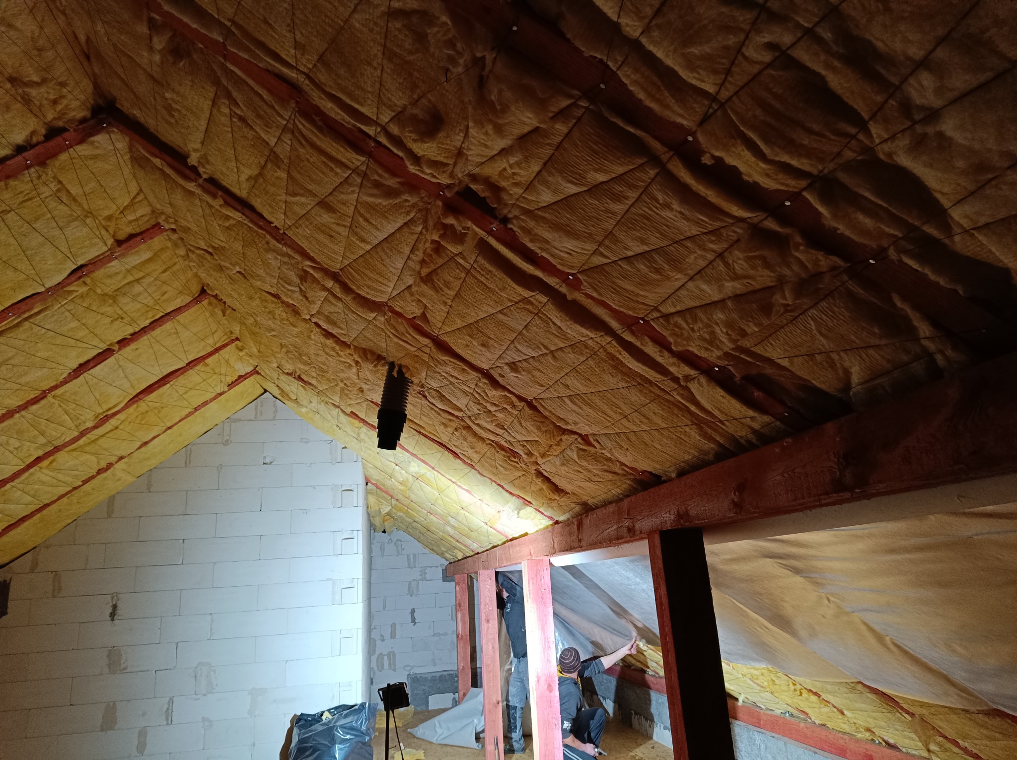 Ocieplenie poddasza wełną mineralną, widoczne drewniane krokwie i pracownicy montujący folię paroizolacyjną, ściana z białych bloczków.
