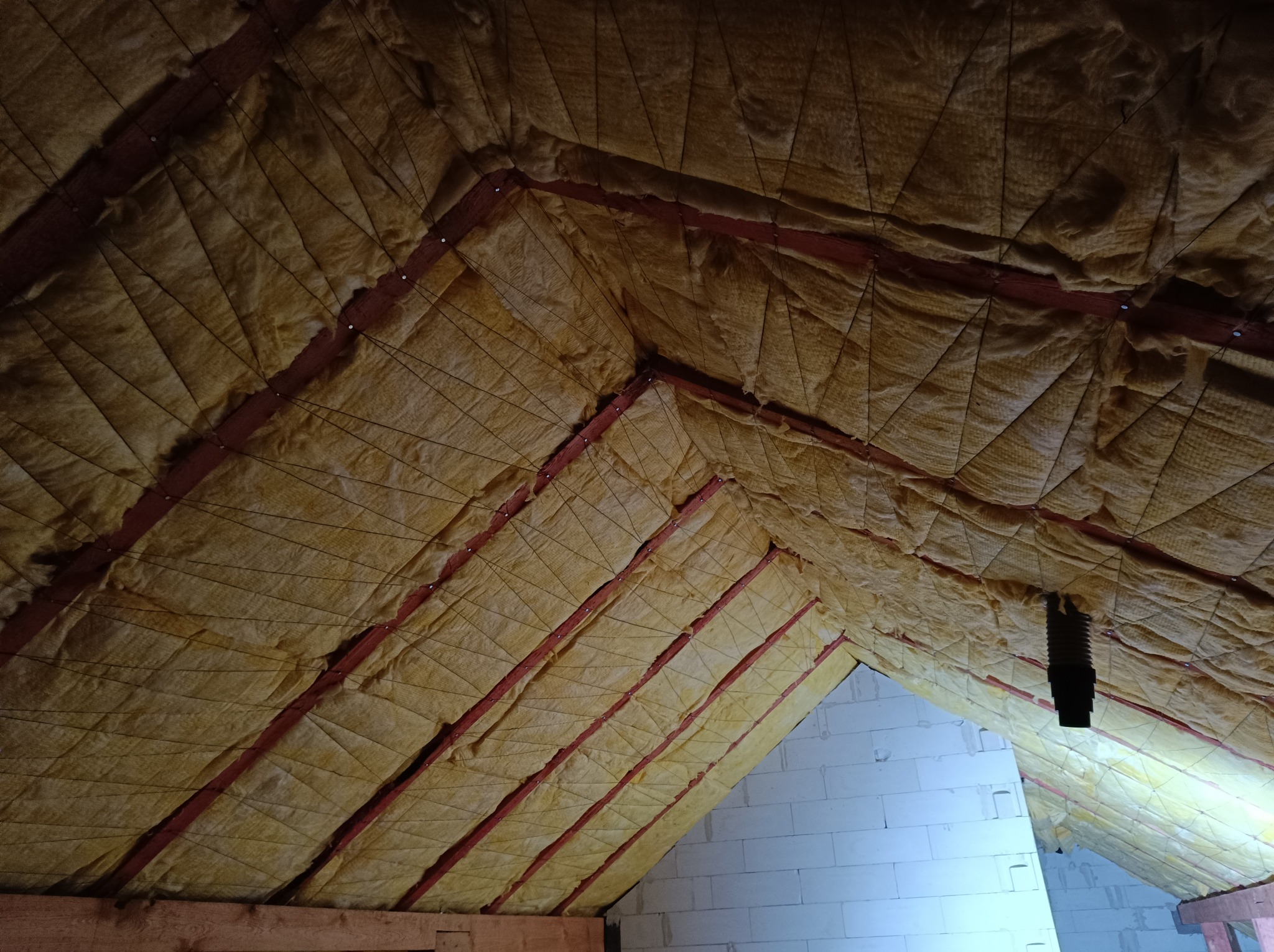 Ocieplenie poddasza wełną mineralną, widoczne krokwie i mocowania, fragment ściany z bloczków betonowych.